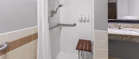 Baño de hotel con ducha, barras de apoyo, un pequeño banco de madera y artículos de tocador en la pared.