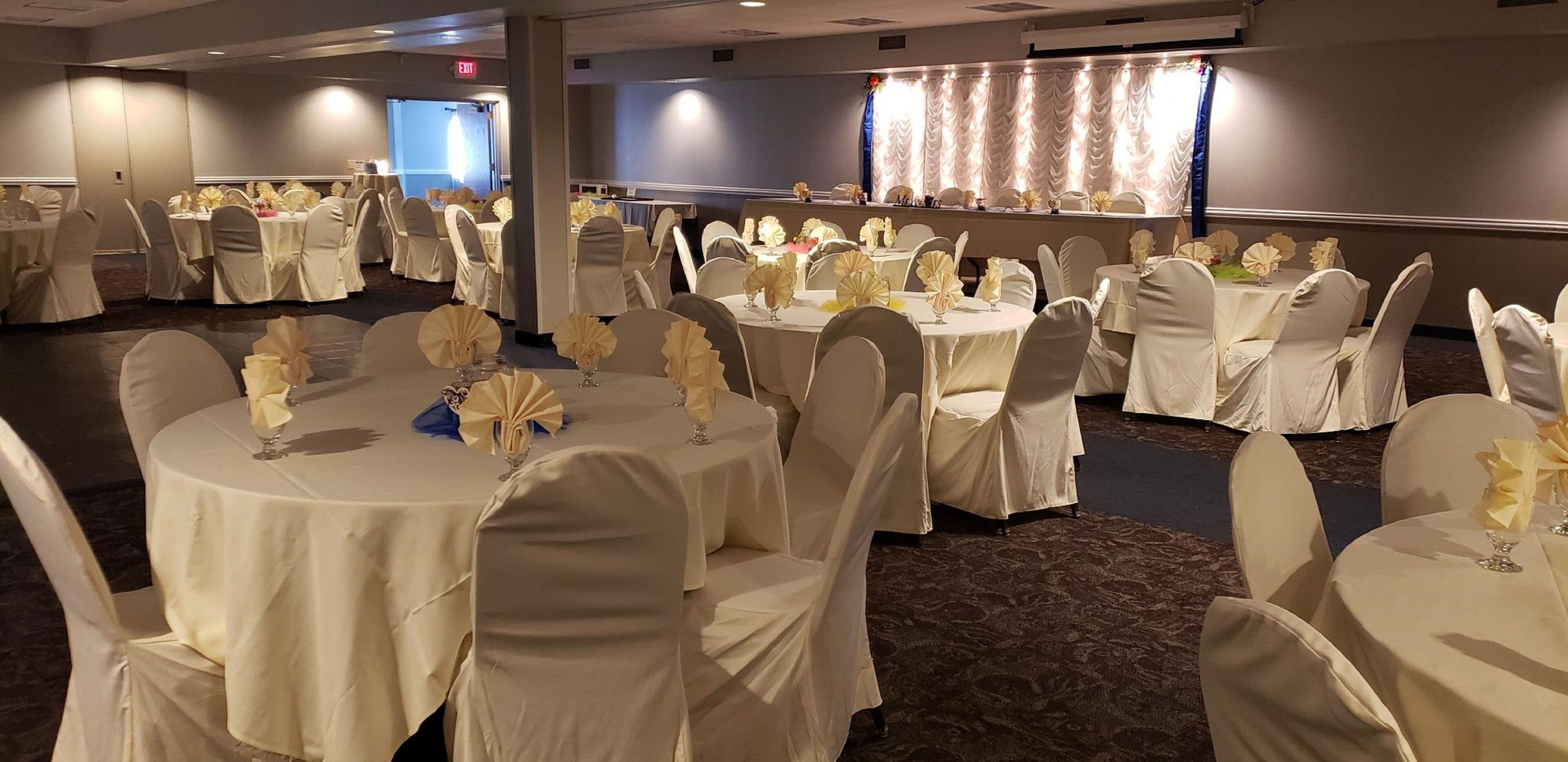 A banquet hall with round tables covered in white tablecloths, each set with white chairs and centerpieces.