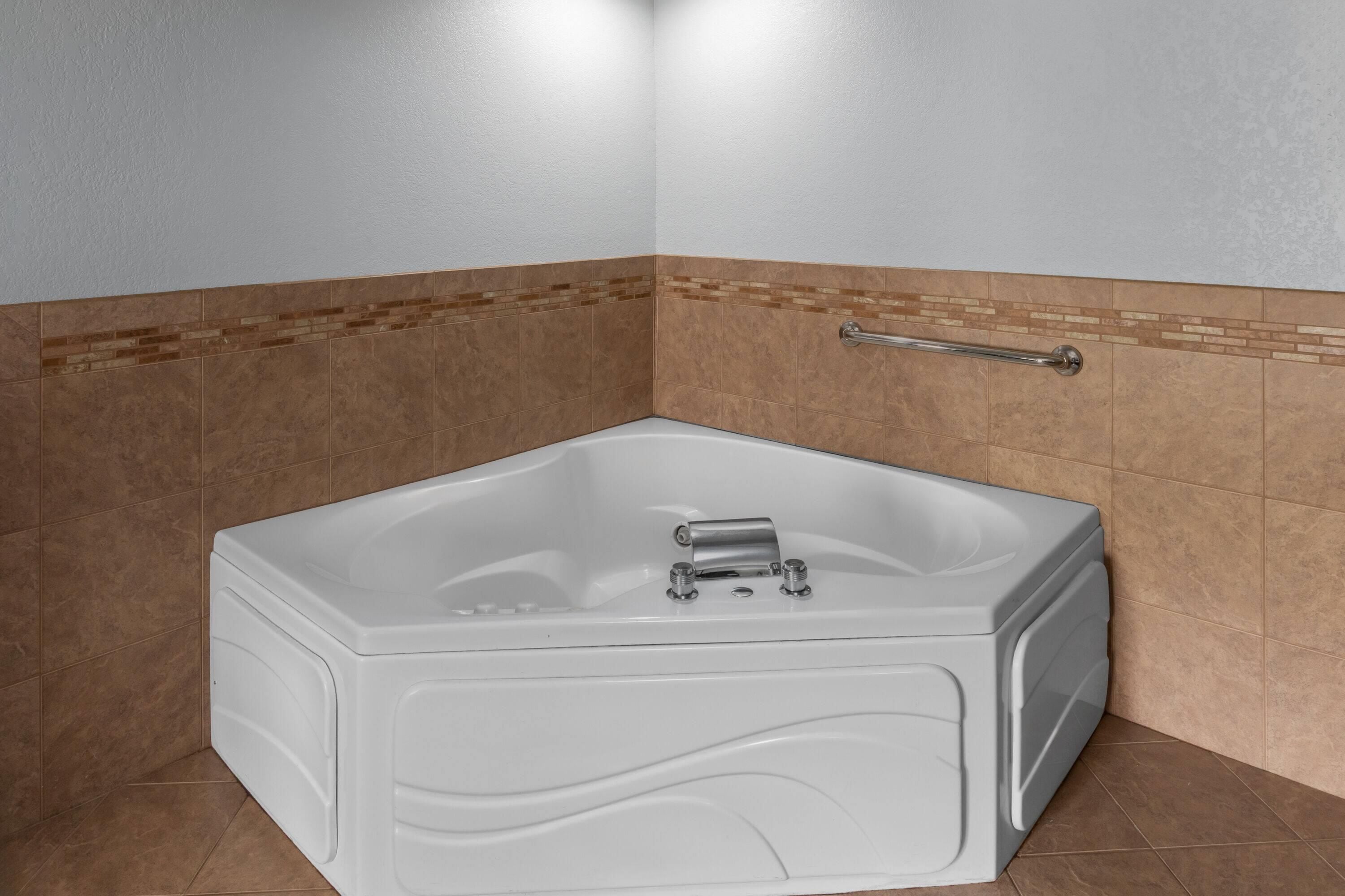 A corner bathroom with a white bathtub, a handrail, and brown tiled walls.