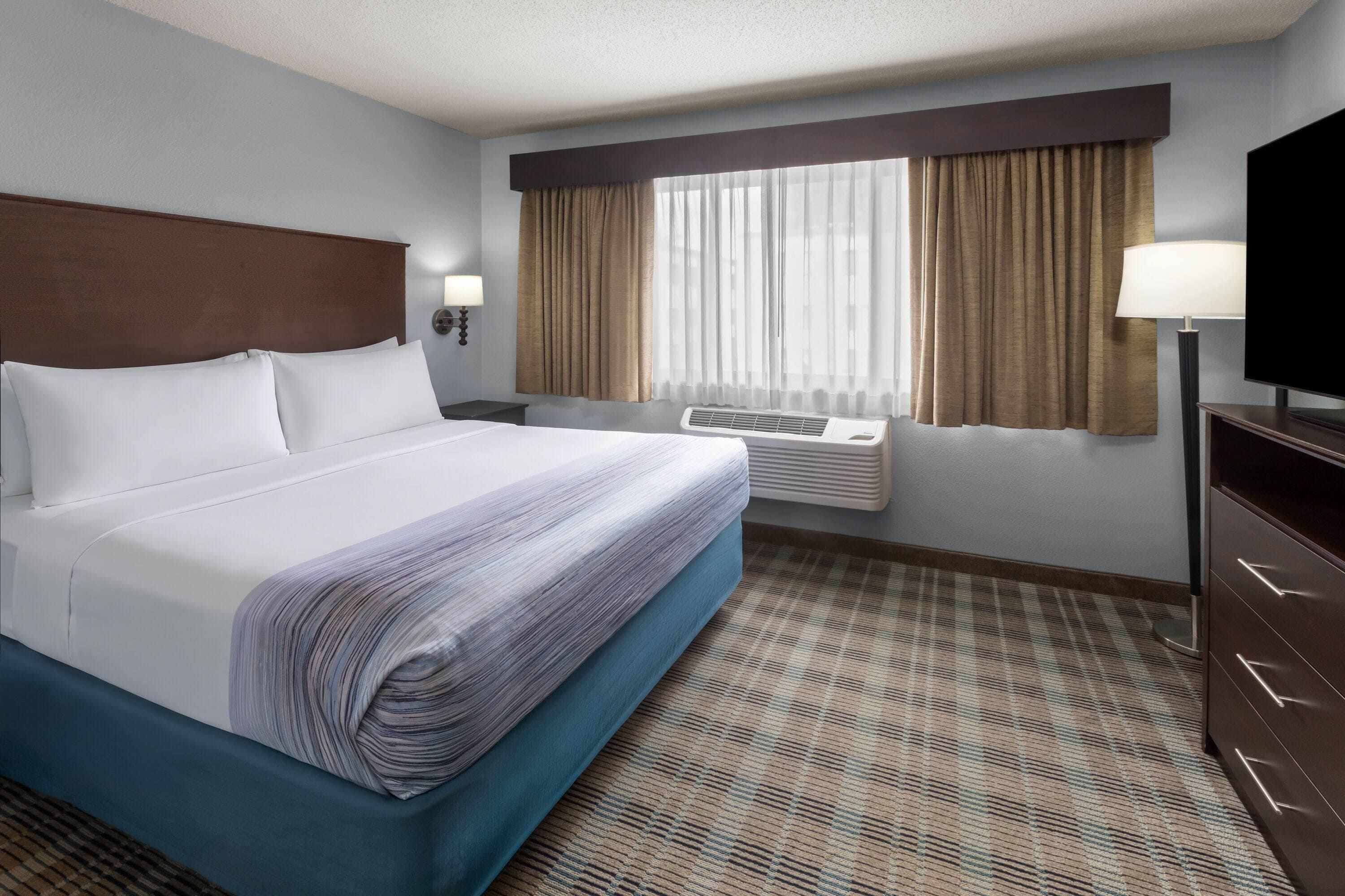 A hotel room with a large bed, a flat-screen TV, a window with curtains, a wall-mounted heater, and a patterned carpet.