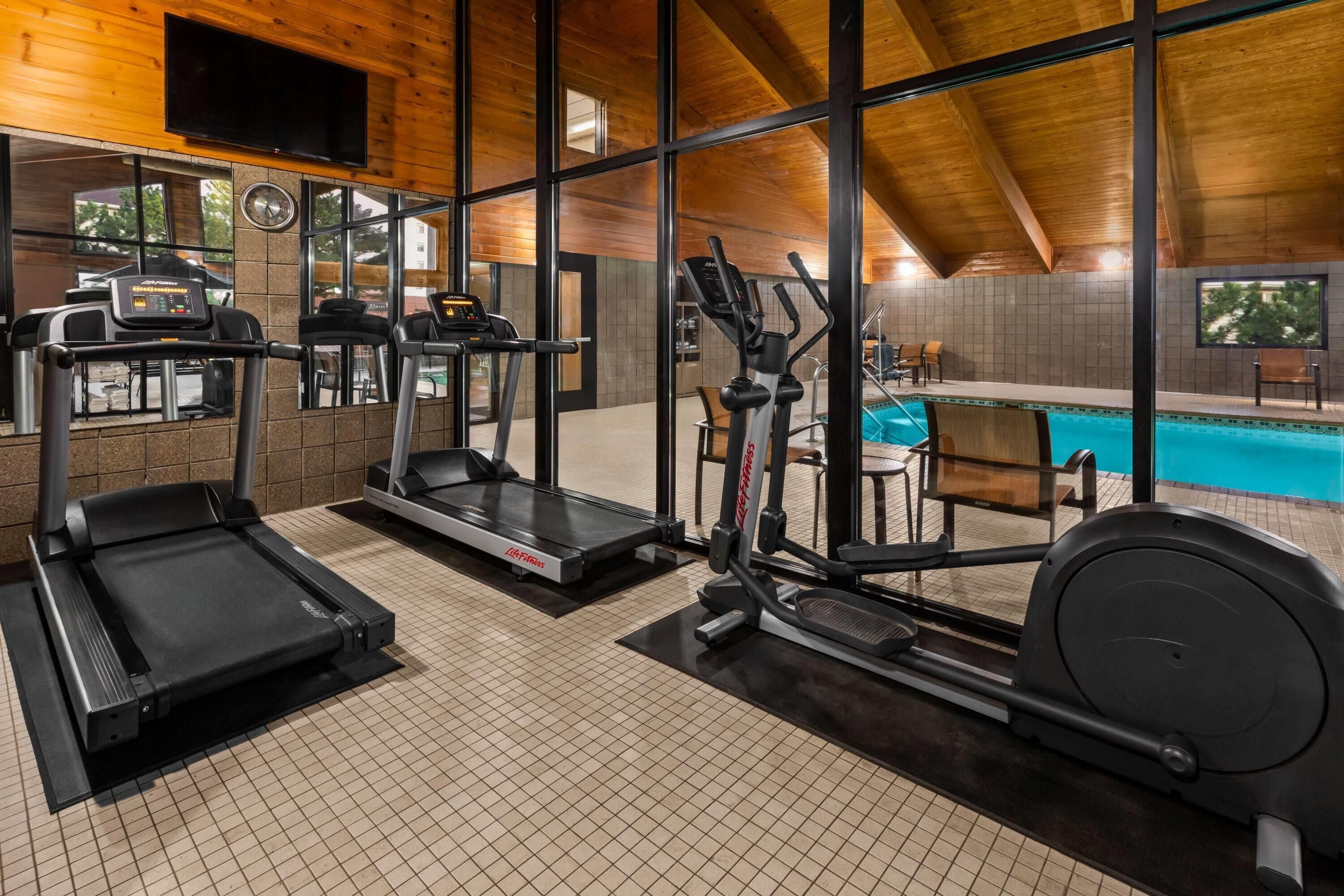 A gym with treadmills, elliptical machines, and a pool area visible through large windows.