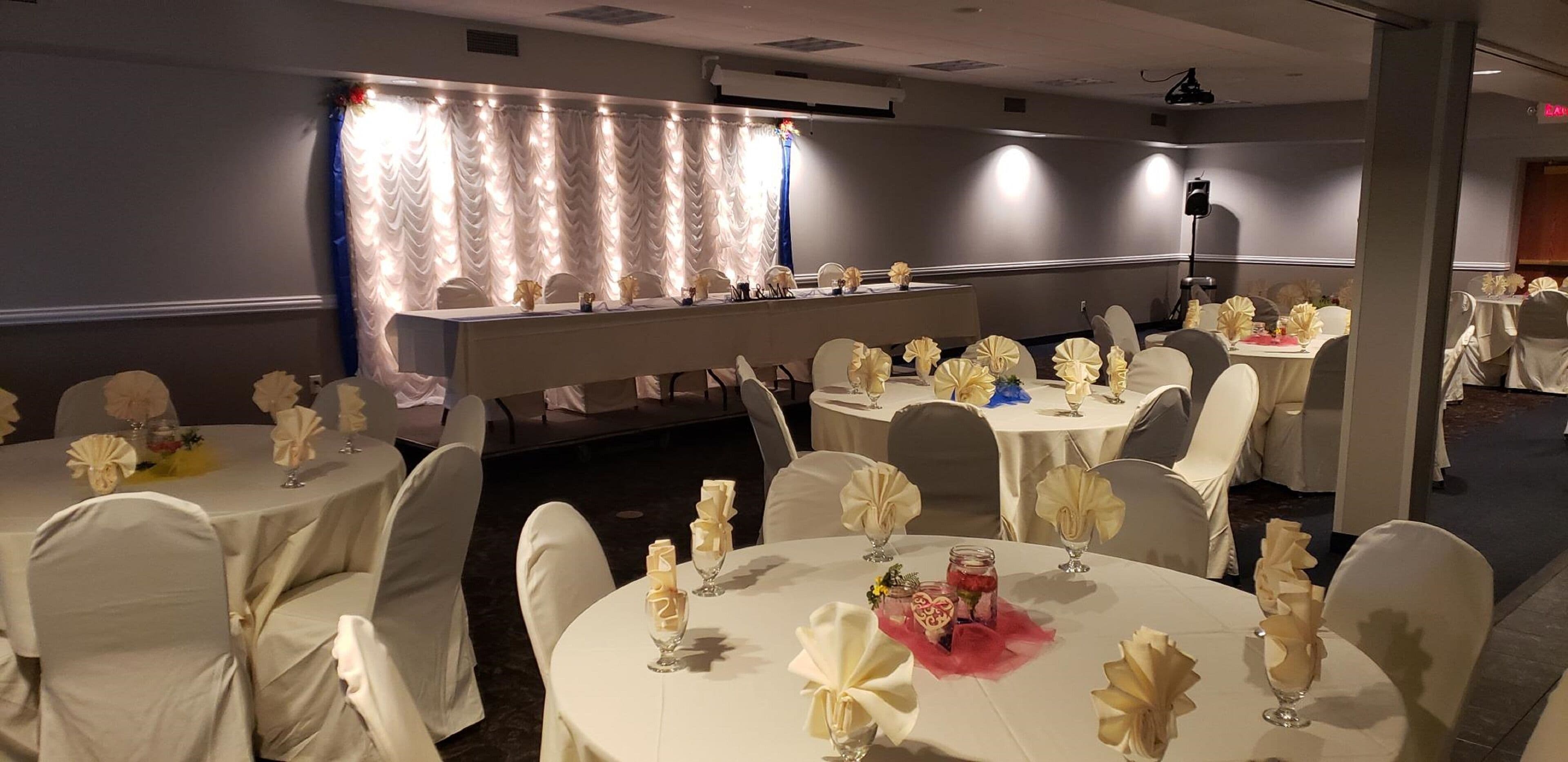 A banquet hall with round tables set for events, featuring white tablecloths and chairs with white covers.