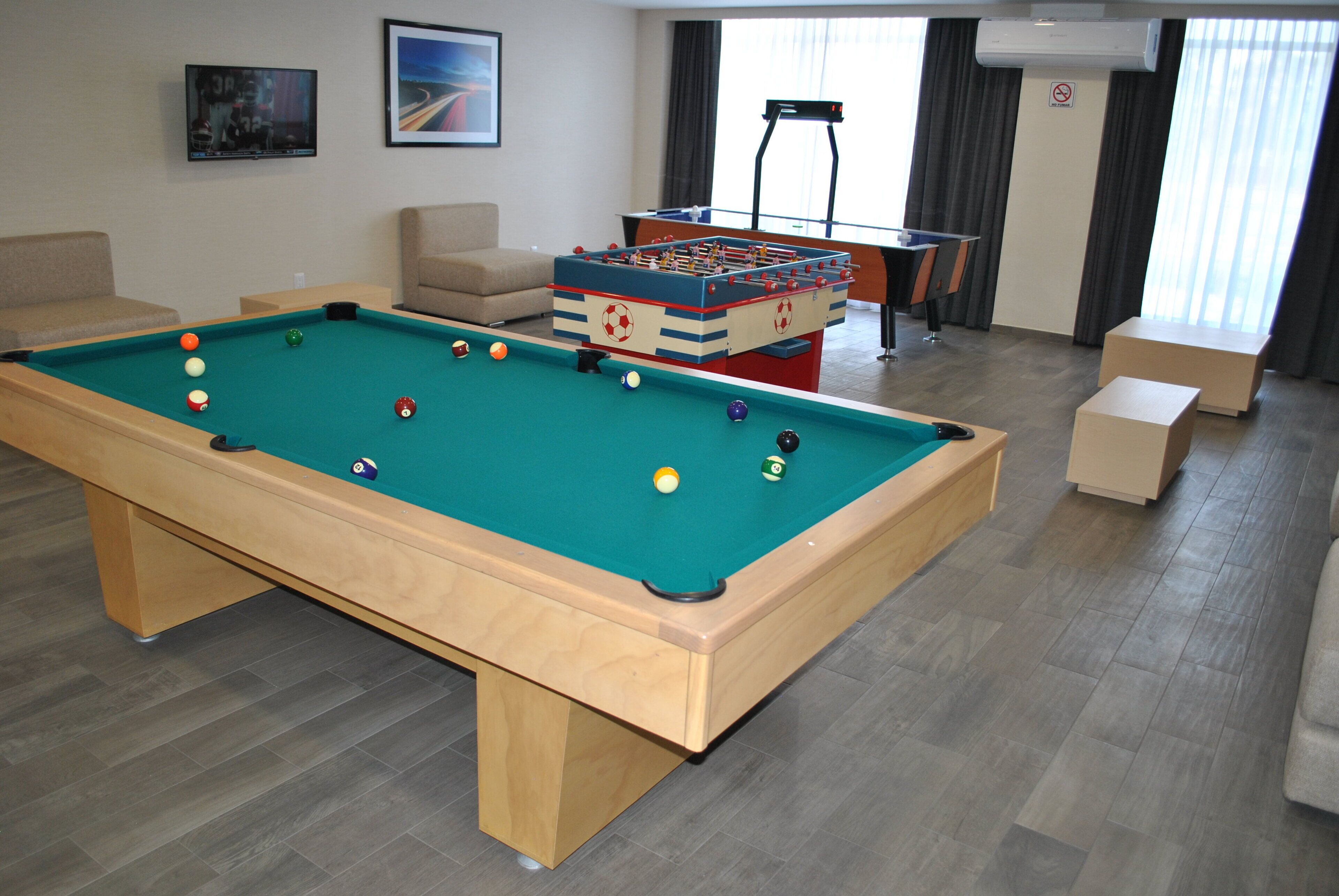 A pool table with balls, a foosball table, a sofa, and a chair in a room with a window and curtains.