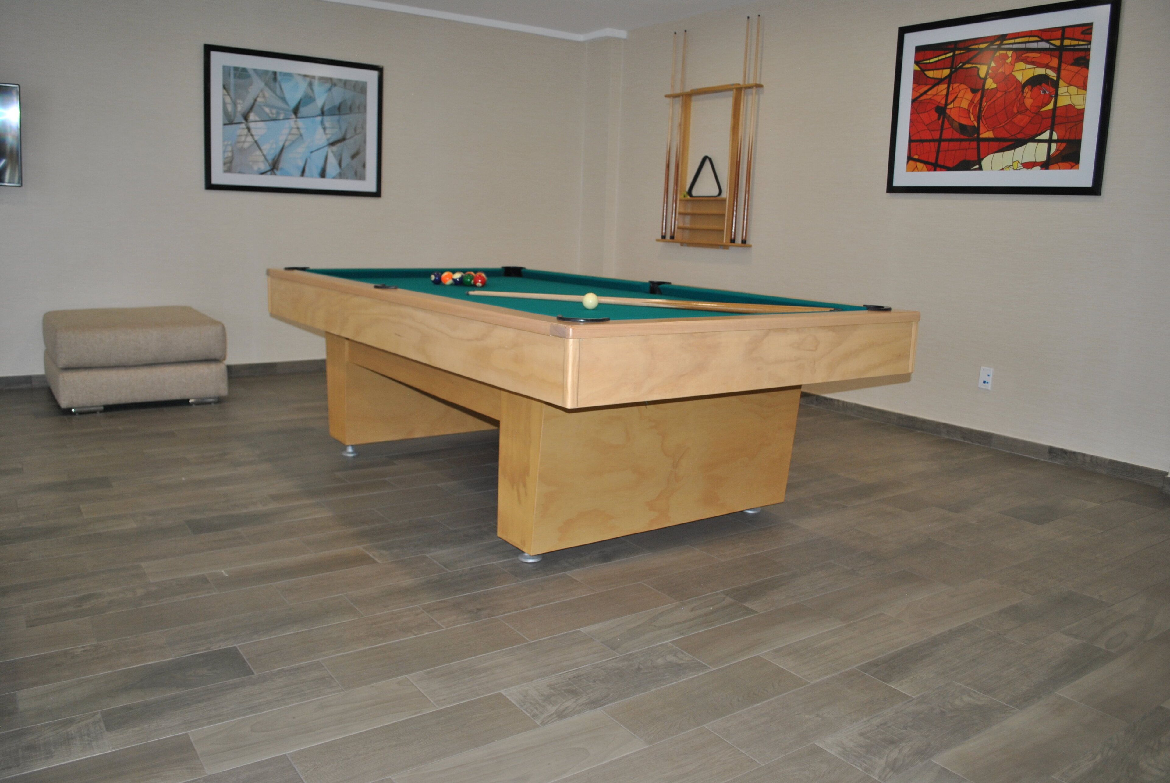 A pool table in a room with wooden flooring, a beige sofa, and two framed artworks on the walls.