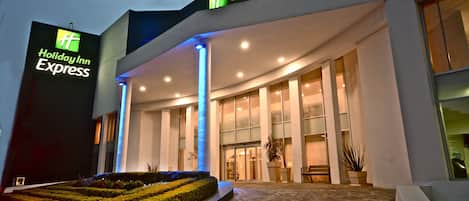 Edificio Holiday Inn Express con fachada moderna, gran entrada de vidrio y zona ajardinada con setos podados.