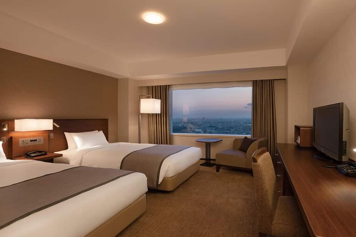 Ein Hotelzimmer mit zwei Betten, einem Schreibtisch mit Fernseher, einem kleinen Tisch und Blick auf eine Stadtlandschaft.