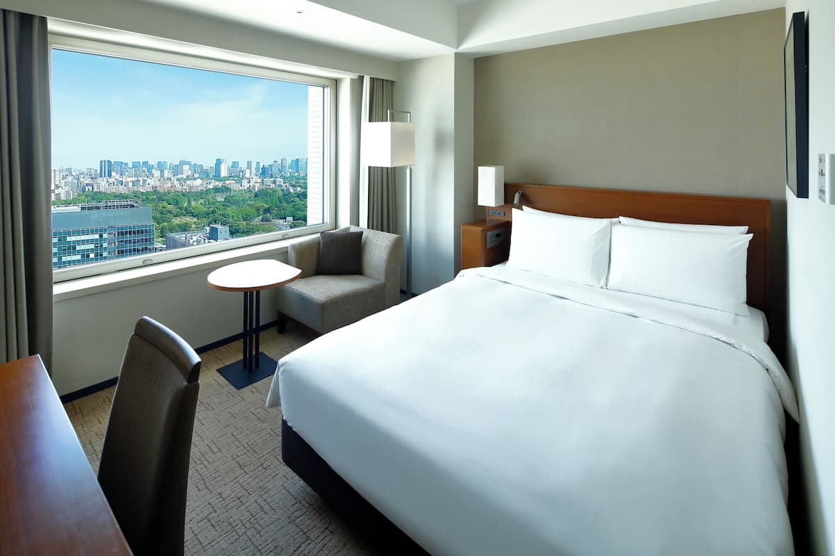 Une chambre d’hôtel avec un grand lit, une petite table, une chaise et une vue sur le paysage urbain, visible à travers une grande fenêtre.