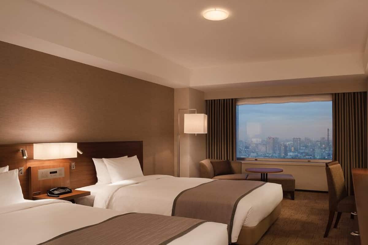 Ein Hotelzimmer mit zwei Betten, einem Schreibtisch, einem Stuhl, einem Fernseher und einem großen Fenster mit Blick auf die Stadt.