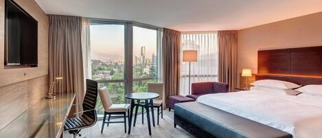 A hotel room with a large bed, a desk with a TV, a chair, a small table, and a view of a cityscape through large windows.