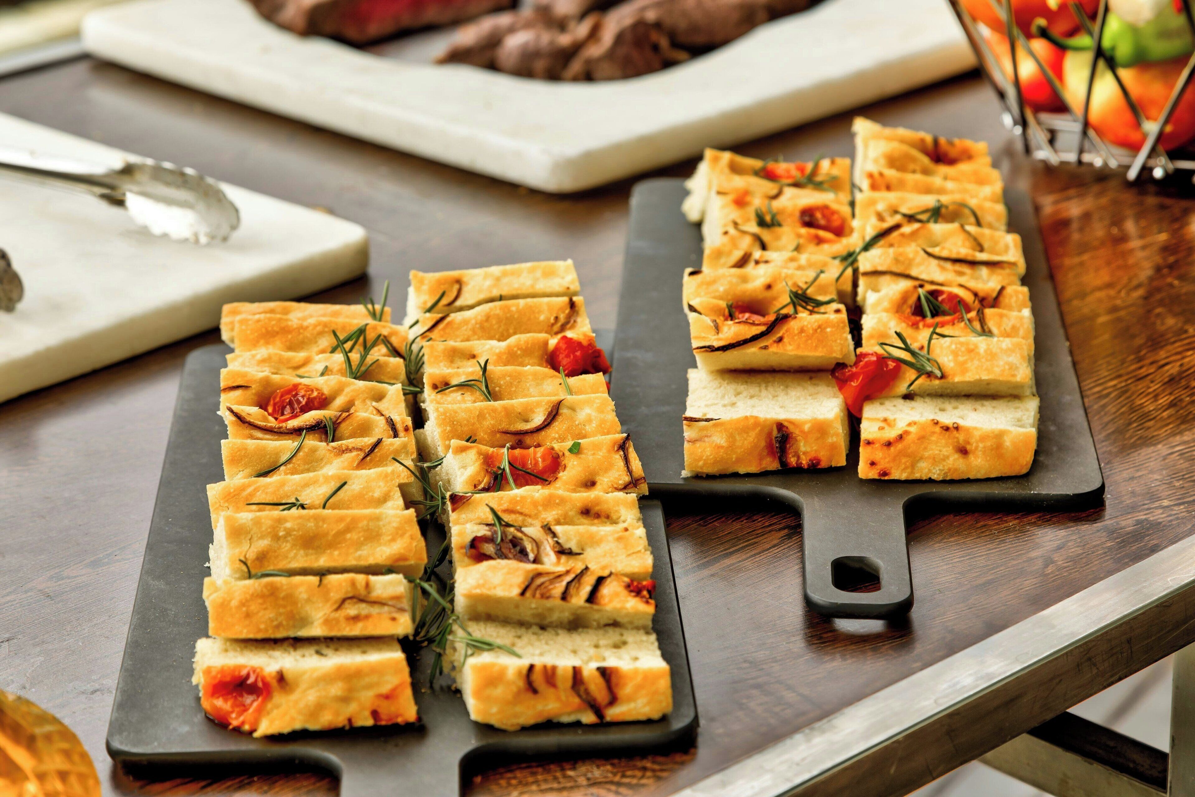 A wooden table with two rectangular serving boards containing neatly arranged square pieces of bread garnished with rosemary and tomatoes.
