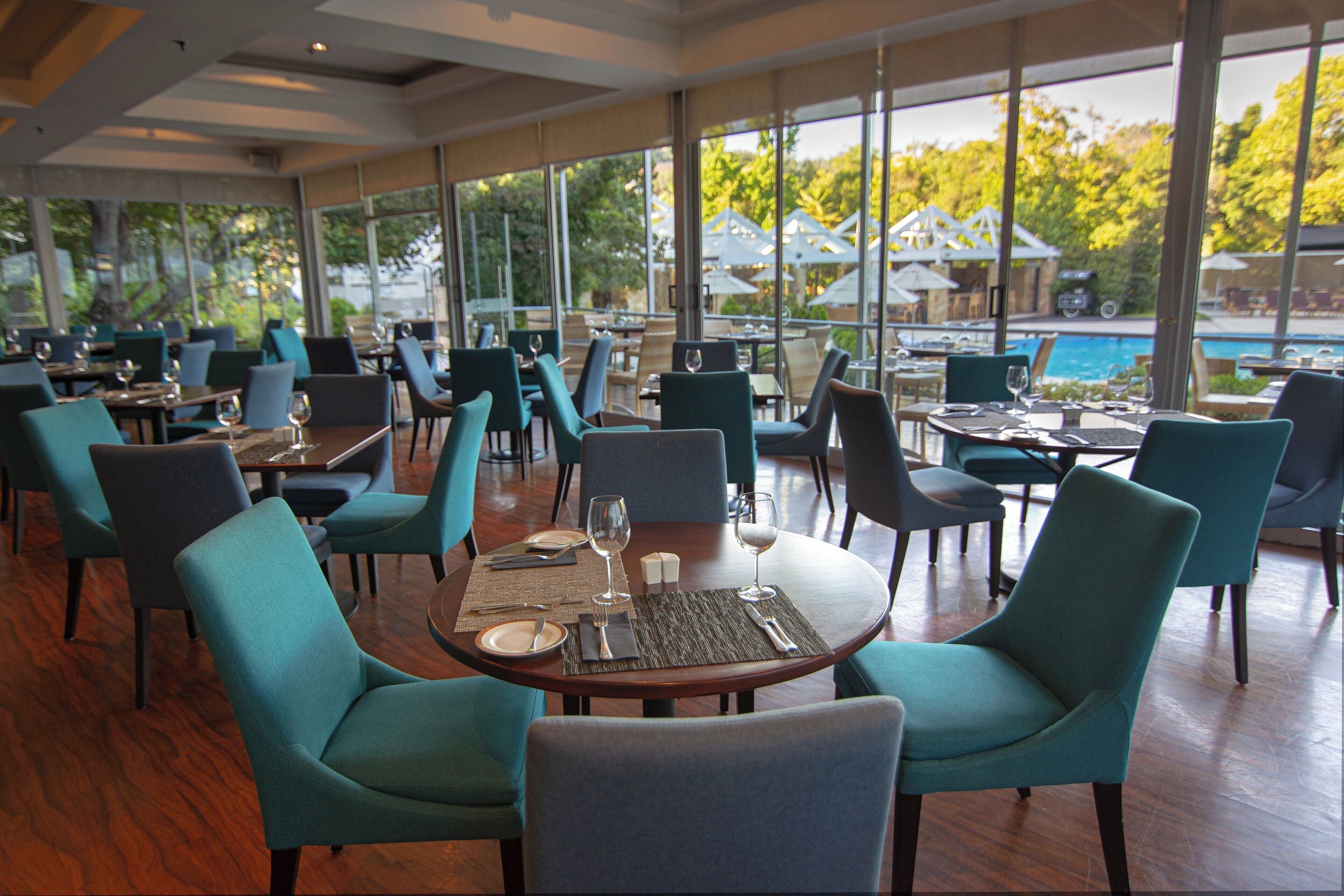 A restaurant with round tables, teal chairs, and a view of a pool and umbrellas outside.