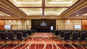 A Sheraton hotel conference room with rows of chairs, a stage, and a large screen displaying the hotel name.
