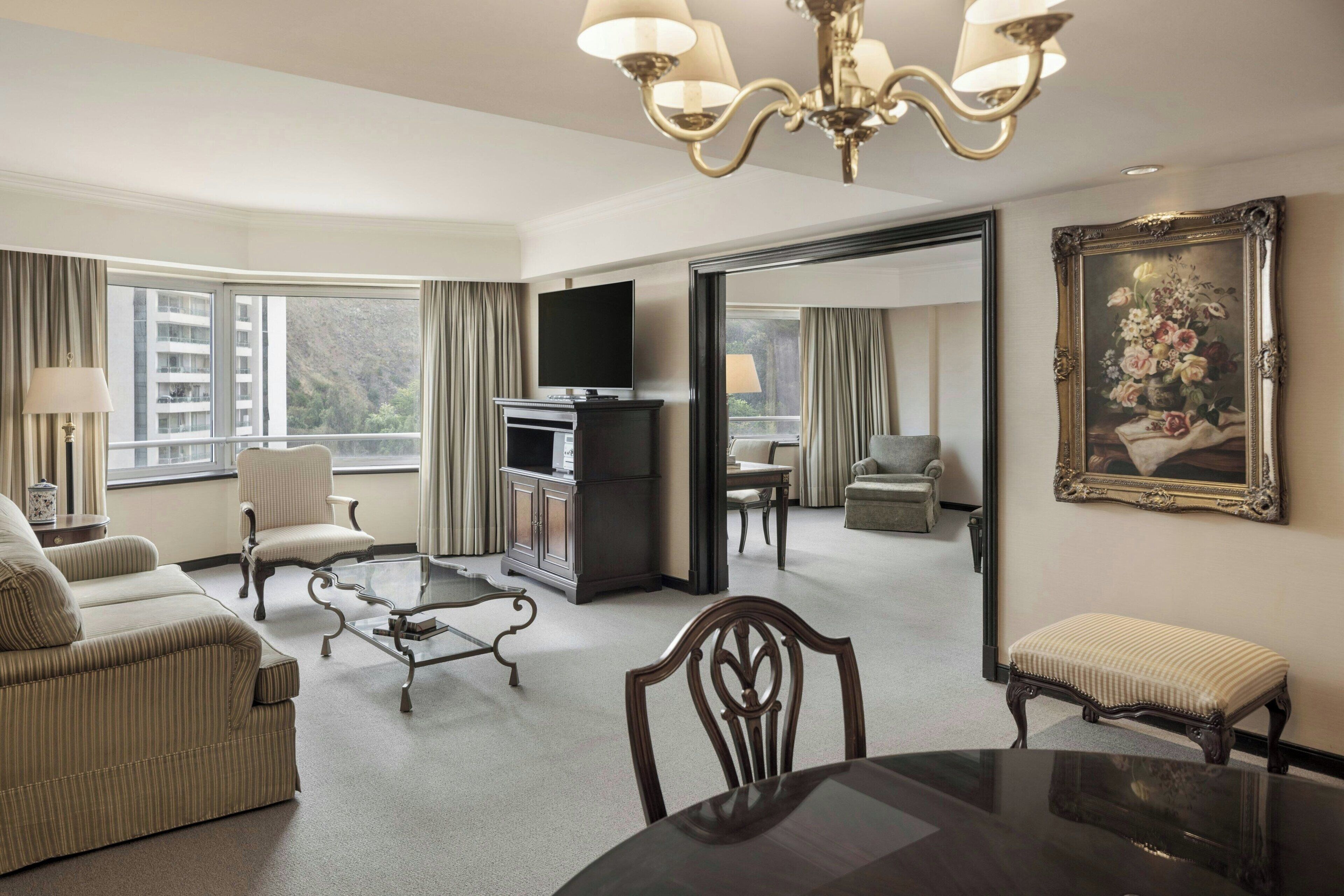 A spacious hotel room with a large window, a flat-screen TV, a dining table, and a sofa.