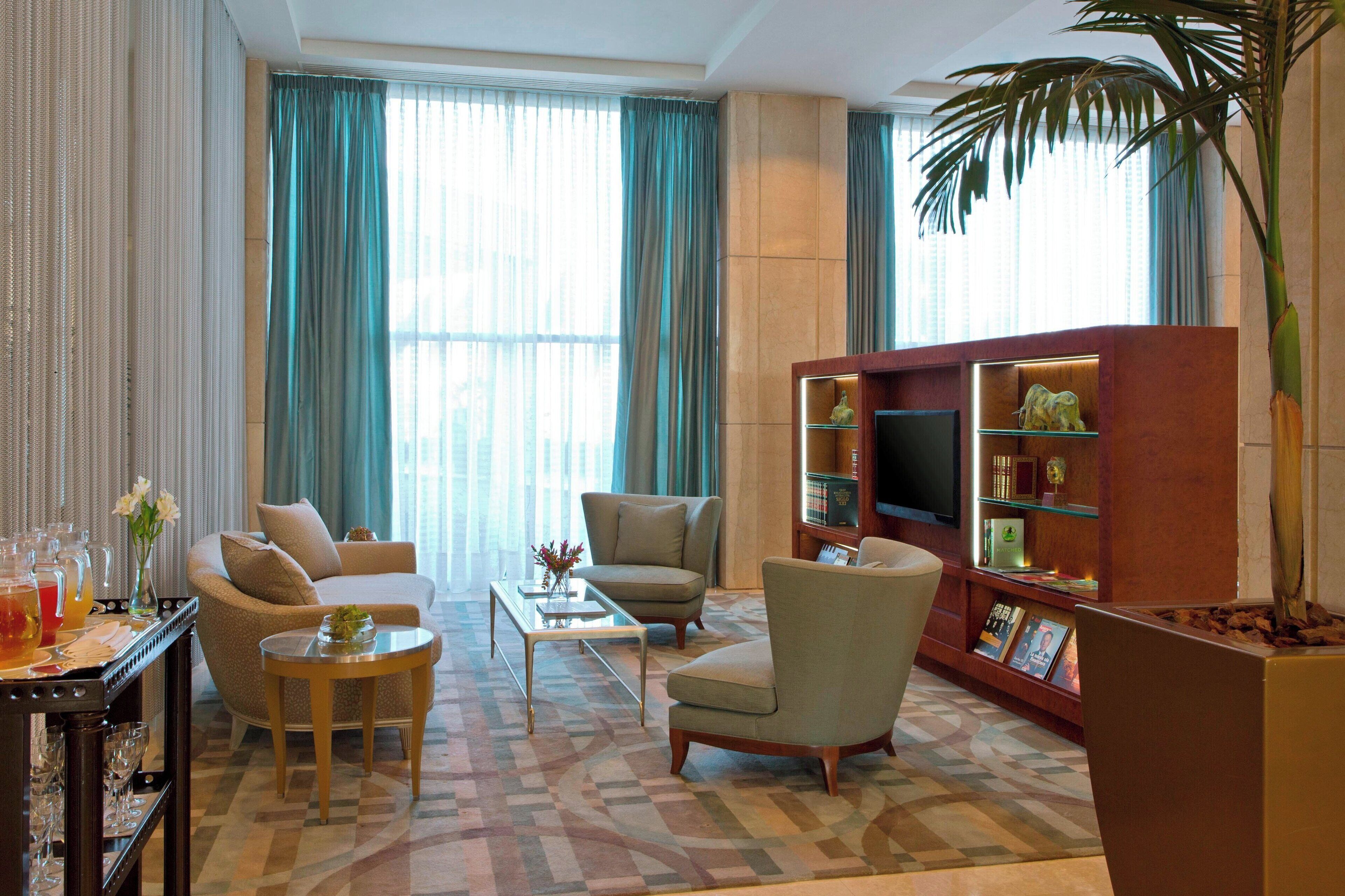 A living room with a sofa, armchairs, a coffee table, a TV stand with books and decorative items, and a large plant.