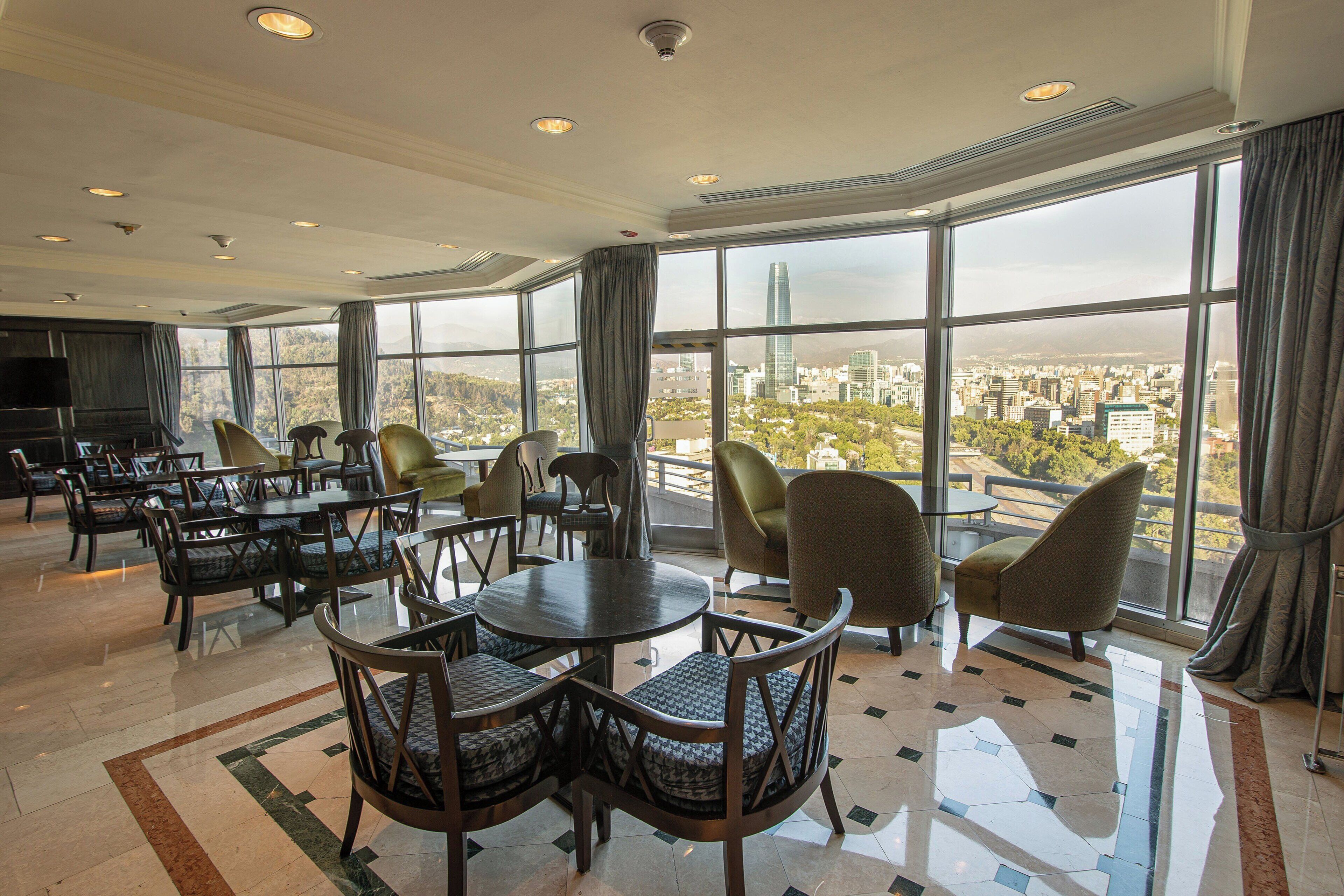 A spacious hotel lobby with a high ceiling, large windows offering a city view, and a variety of seating arrangements including tables and chairs.