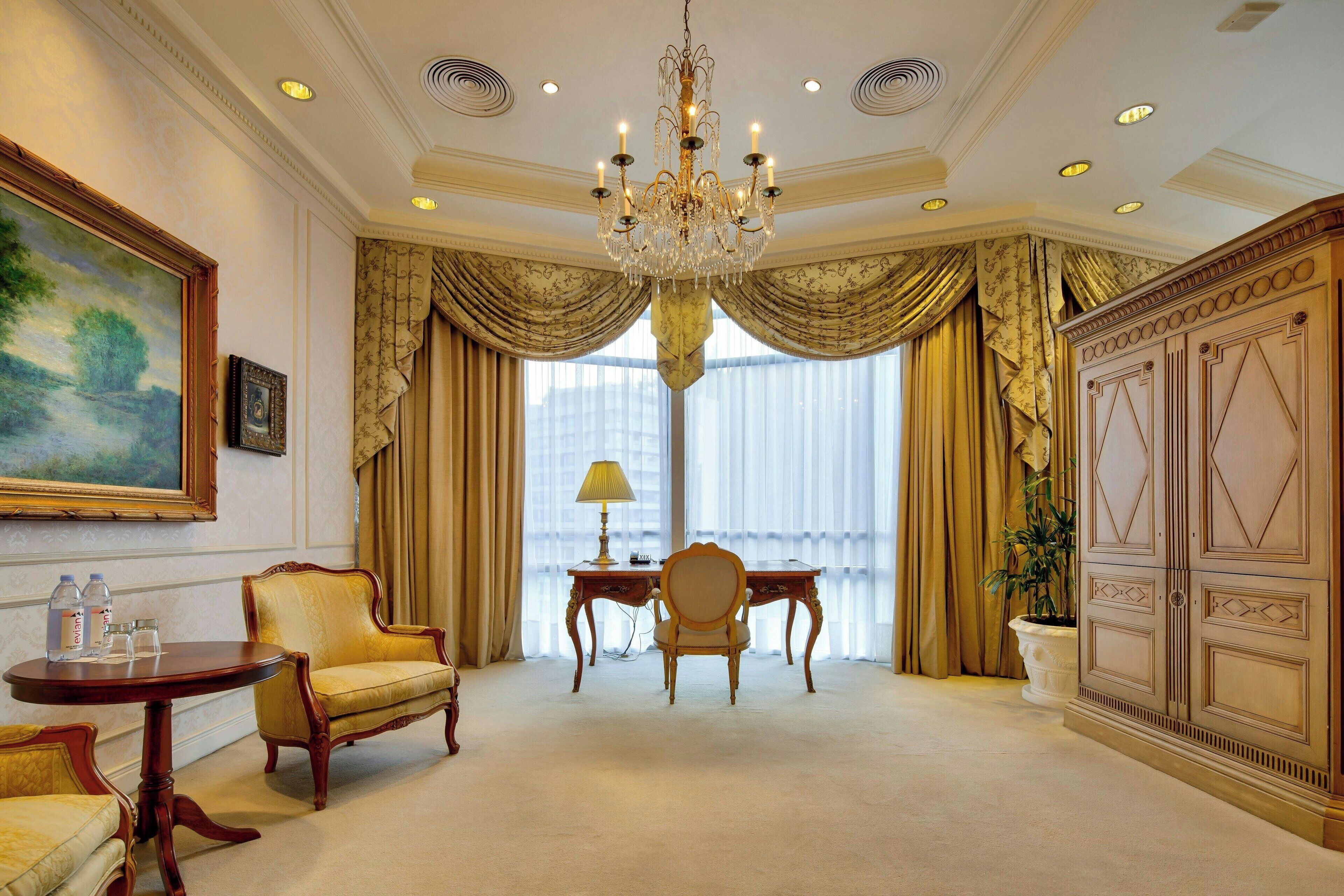 A spacious room with a chandelier, a painting, a desk with a lamp, and a chair.
