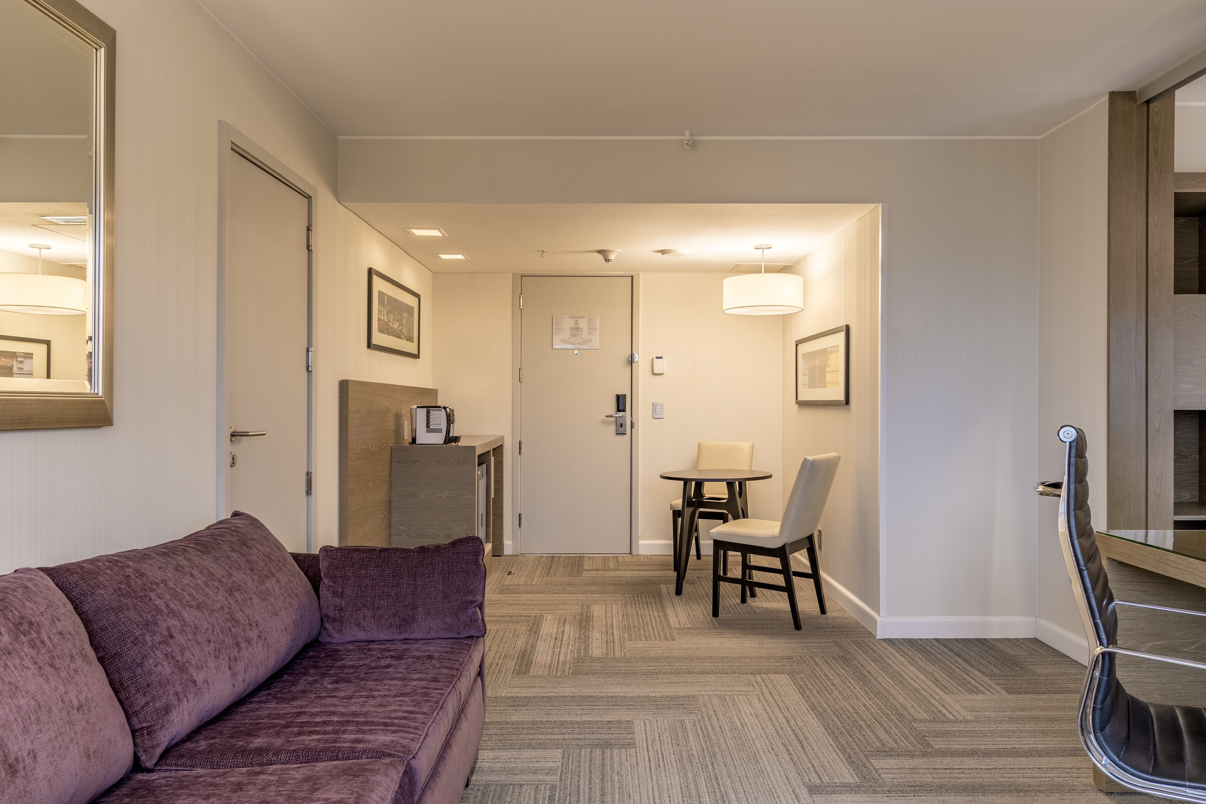 Una habitación de hotel moderna con un sofá morado, un área de comedor con una mesa y sillas, y una escalera de madera.