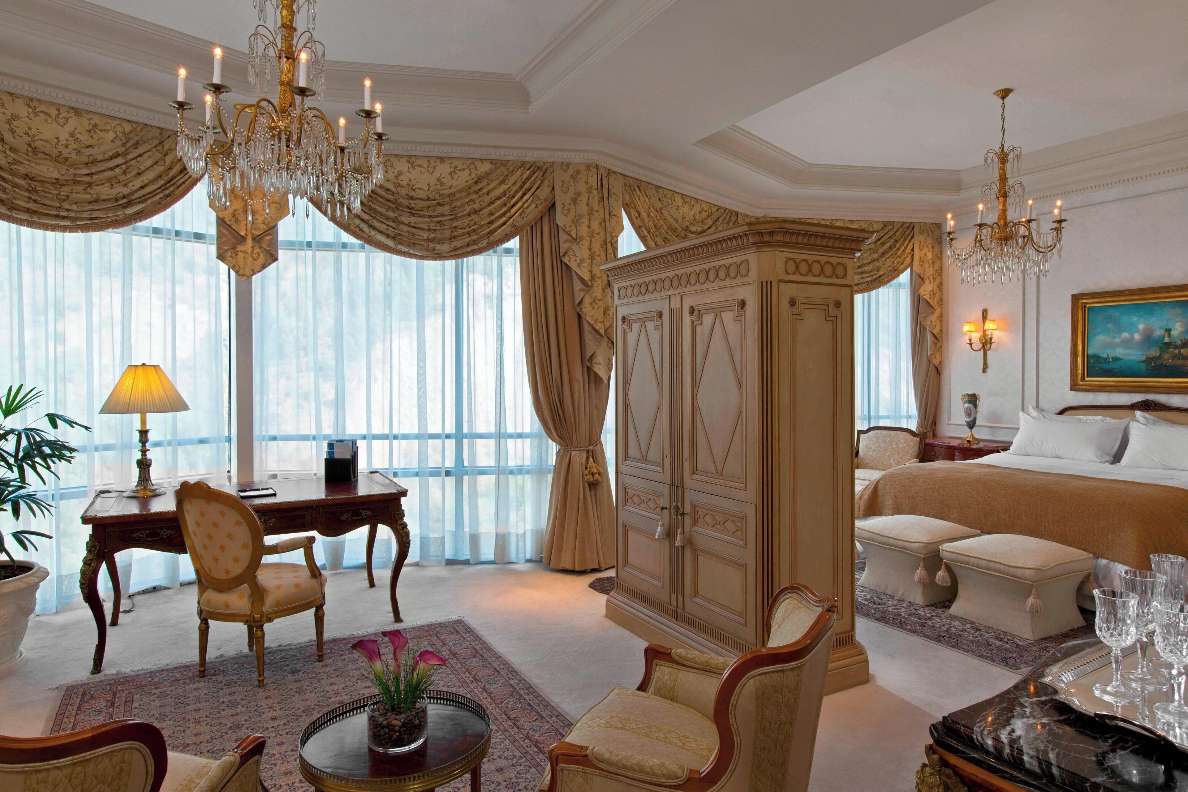 Quarto de hotel luxuoso com uma cama grande, área de estar com escrivaninha e cadeiras, lustre e uma janela ampla com cortinas.