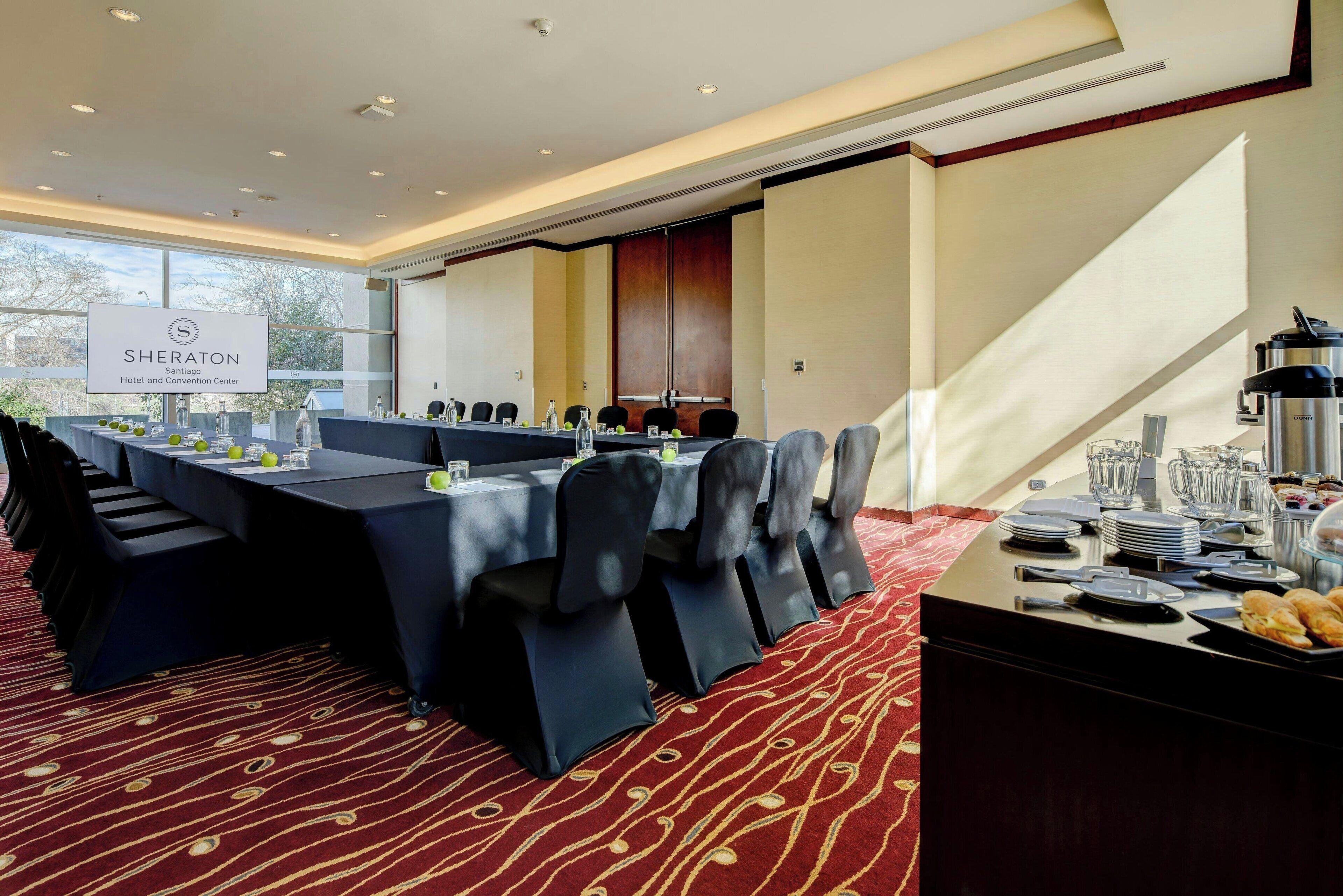 Un salón de conferencias con una larga mesa, sillas y una zona de buffet con platos y vasos.