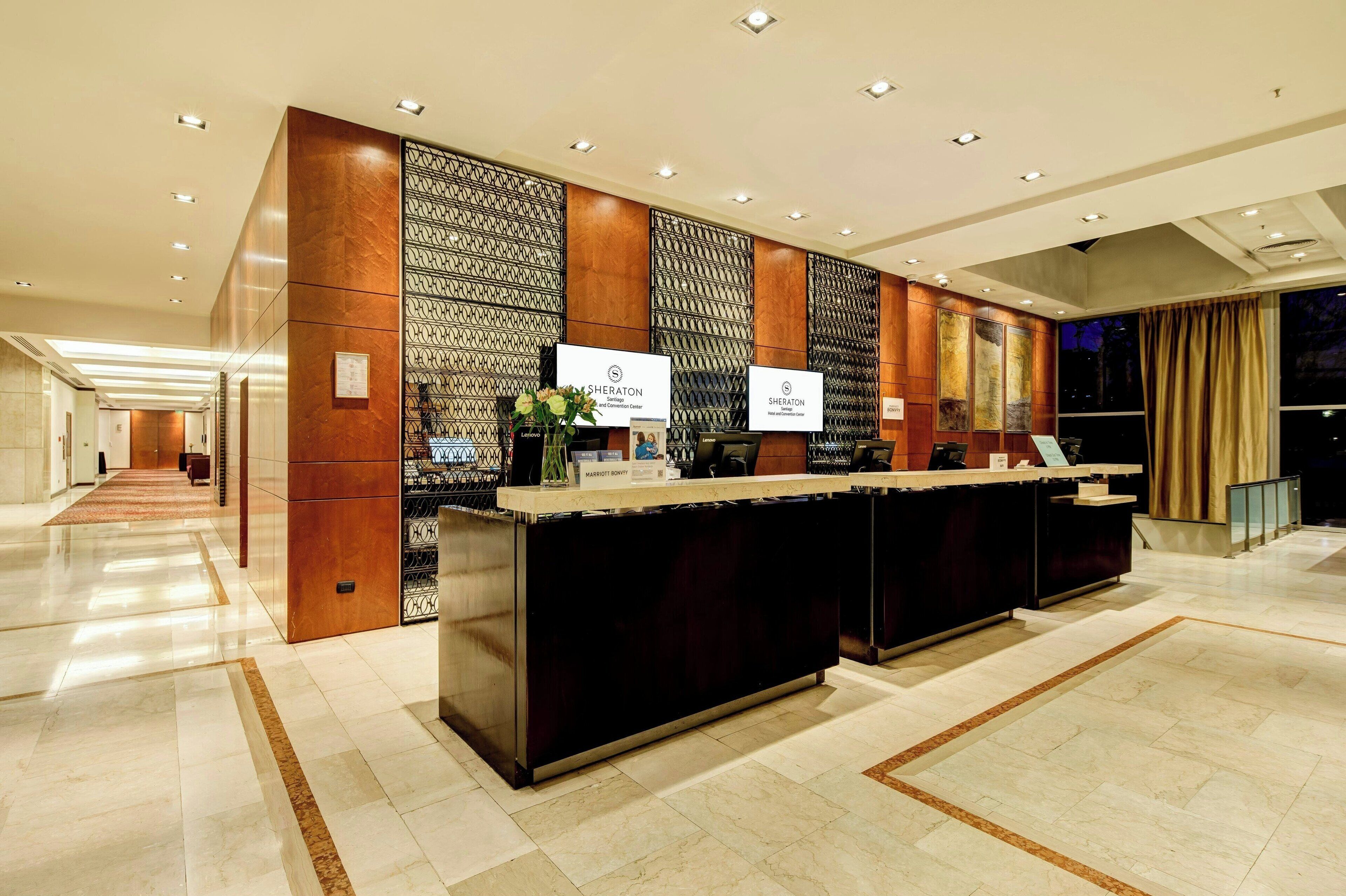 A hotel reception area with a modern design, featuring a reception desk, a wall-mounted information board, and a large window with curtains.