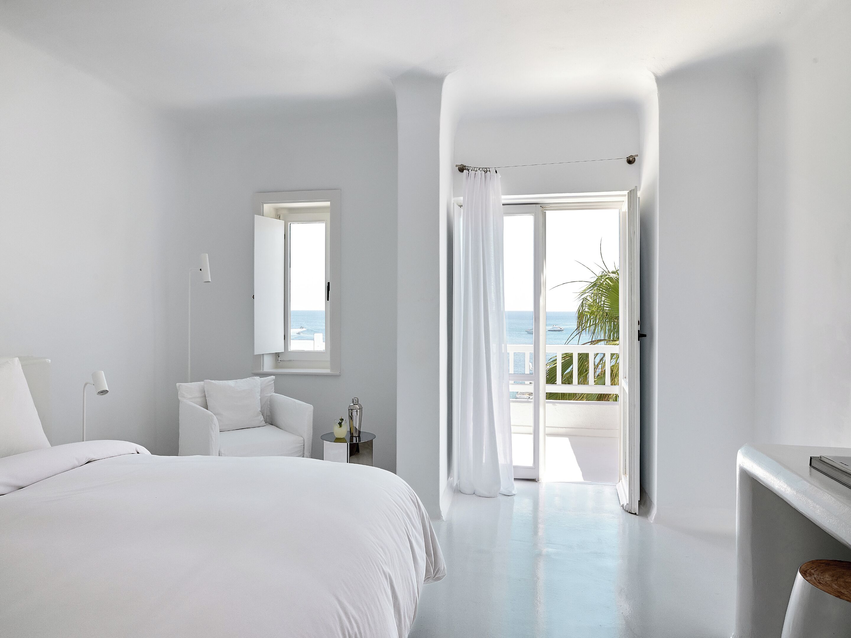 A minimalist bedroom with a large bed, a bedside table, a mirror, and a view of the sea through an open door.
