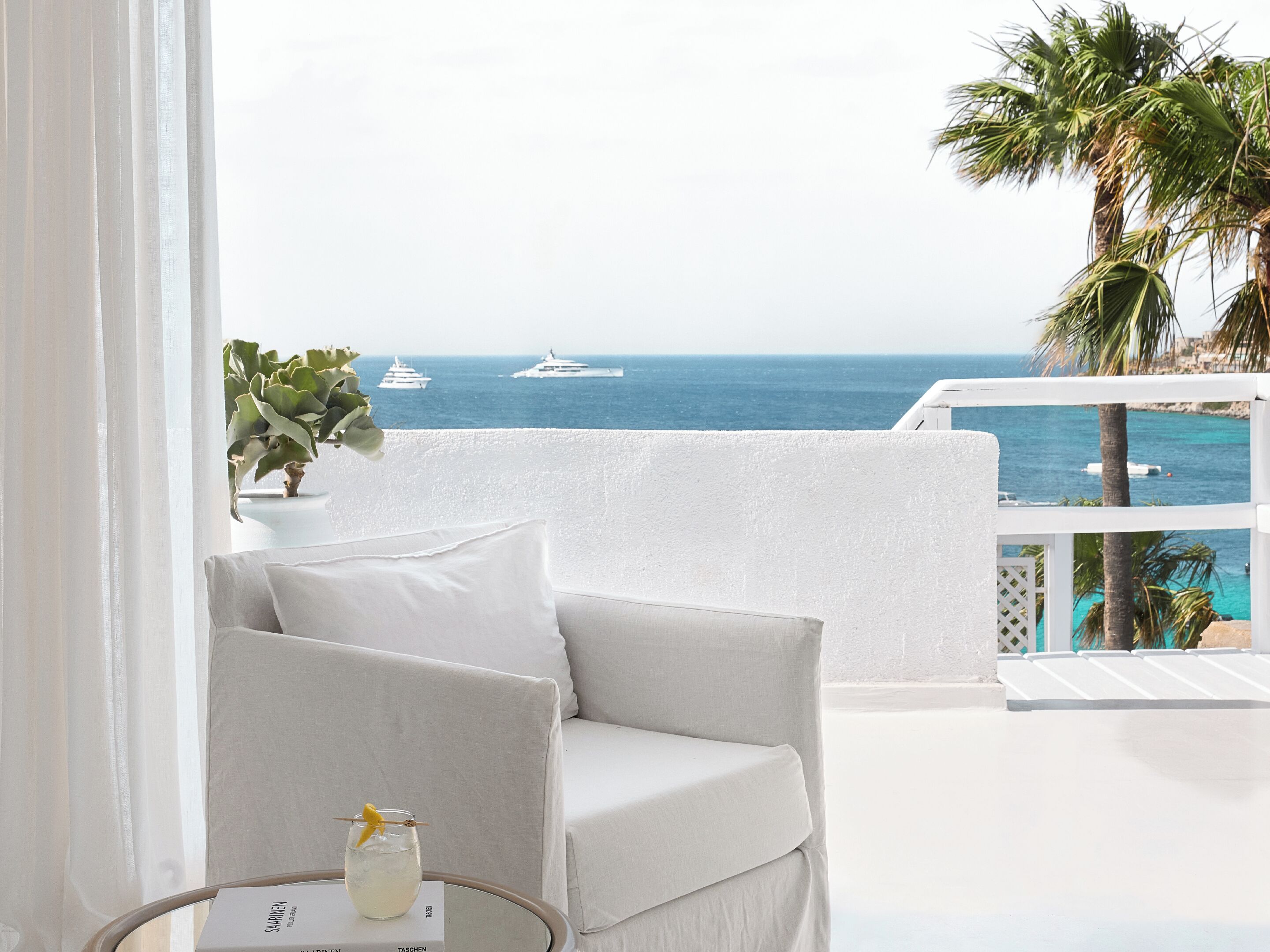 A balcony with a white sofa, a glass table with a drink, and a view of the sea with yachts.