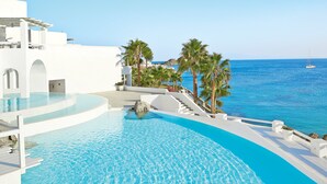 A pool with a curved edge, a white building with arches, and a view of the sea with a sailboat.