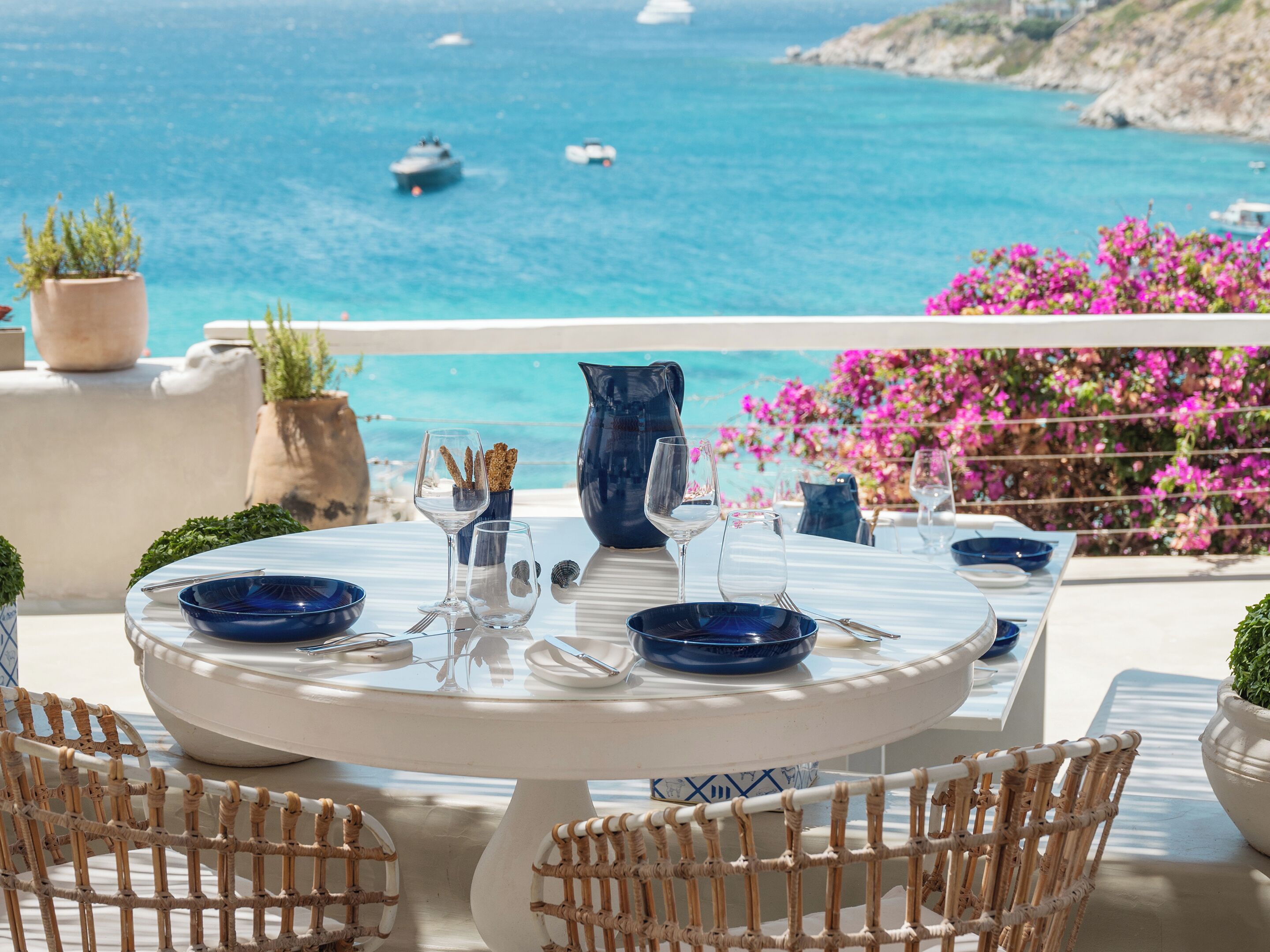 A round table with blue and white dinnerware, surrounded by wicker chairs, overlooking a coastal view with boats and pink flowers.