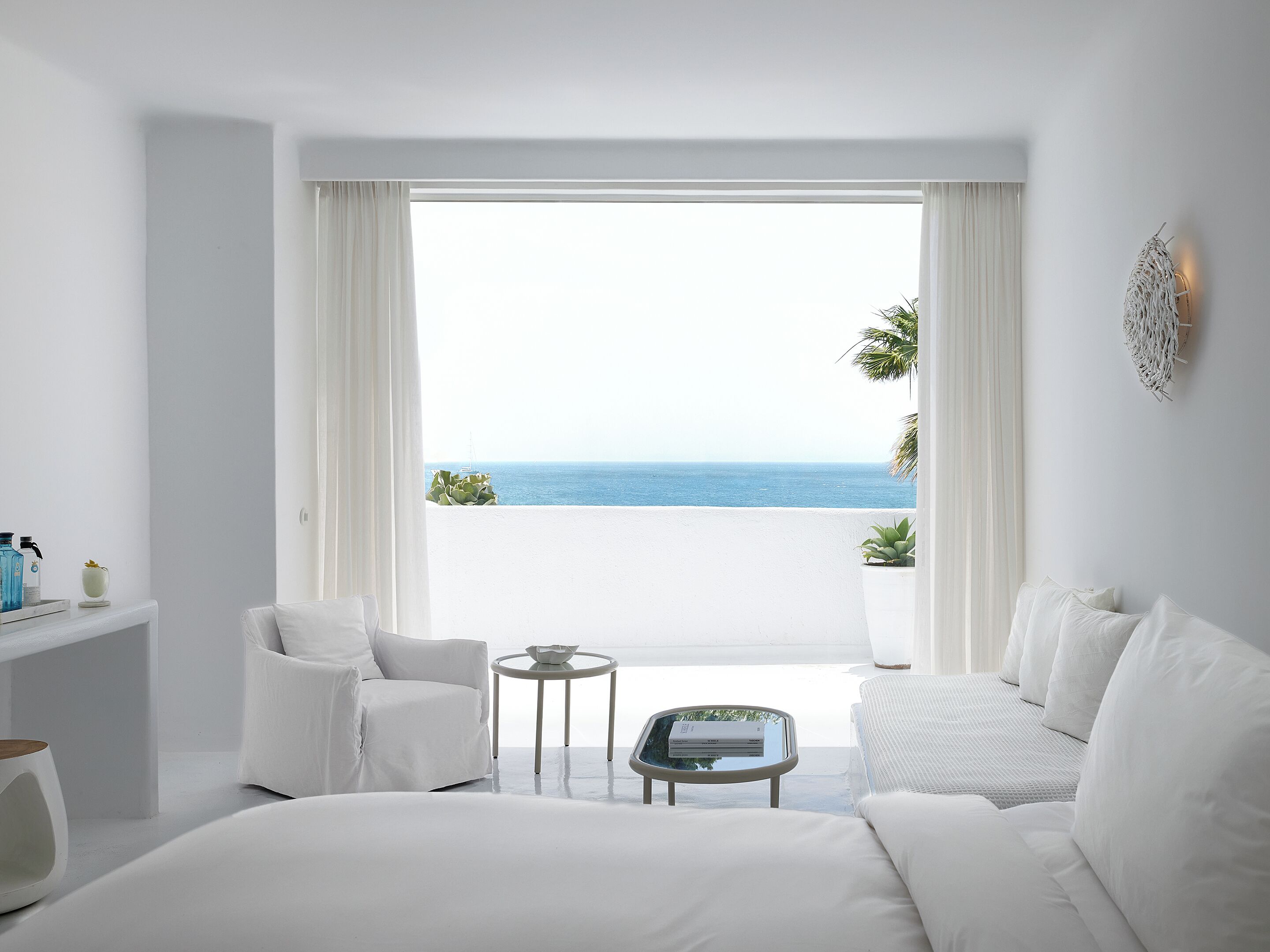 A modern hotel room with a large bed, a white armchair, a small round table, and a view of the sea through large windows.