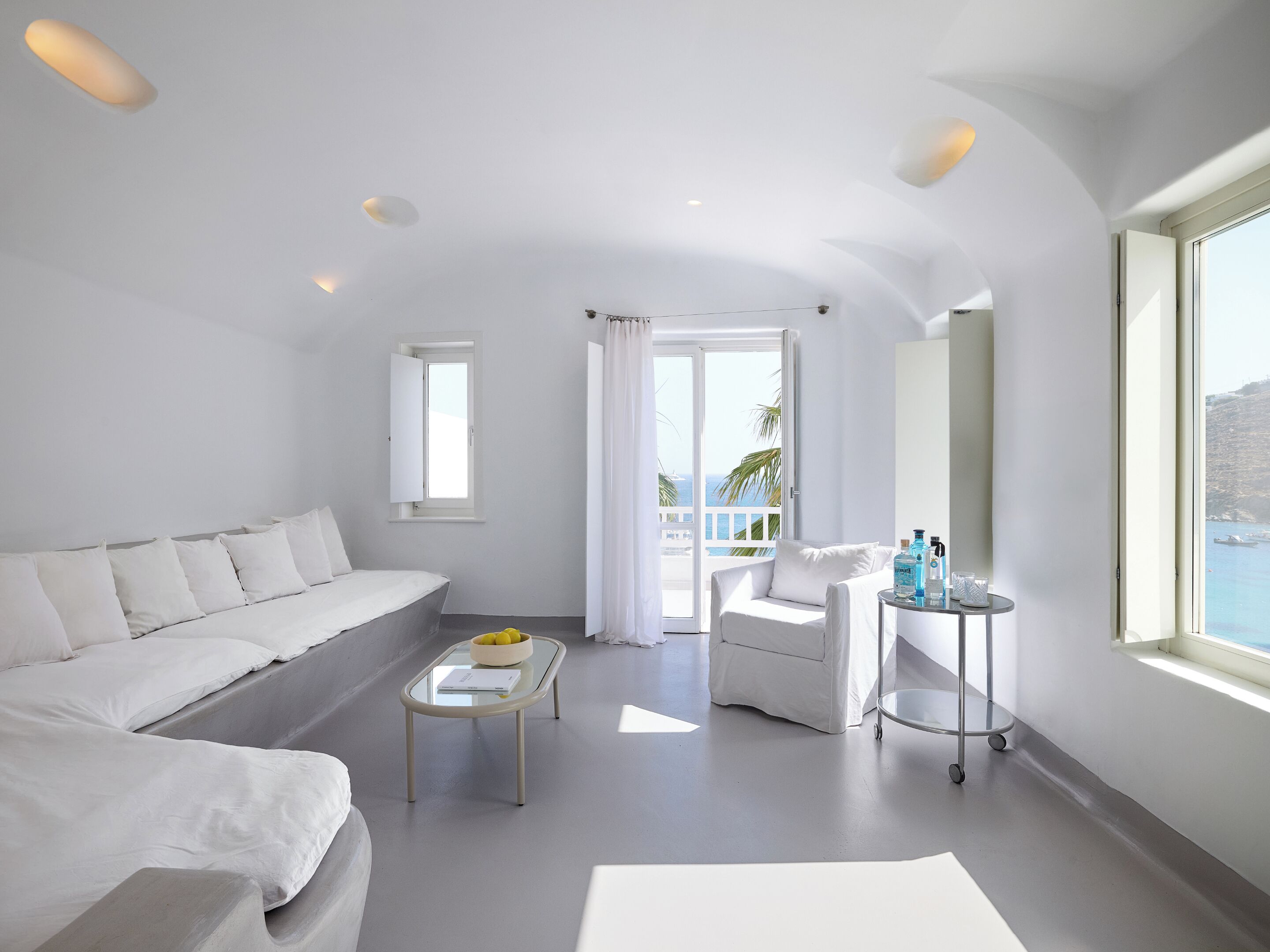 Una habitación moderna y minimalista con un sofá blanco, una mesita redonda y una silla. Un ventanal grande ofrece vistas al mar.