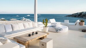 A sunlit outdoor seating area with white furniture, a wooden coffee table, and a view of the sea with yachts.