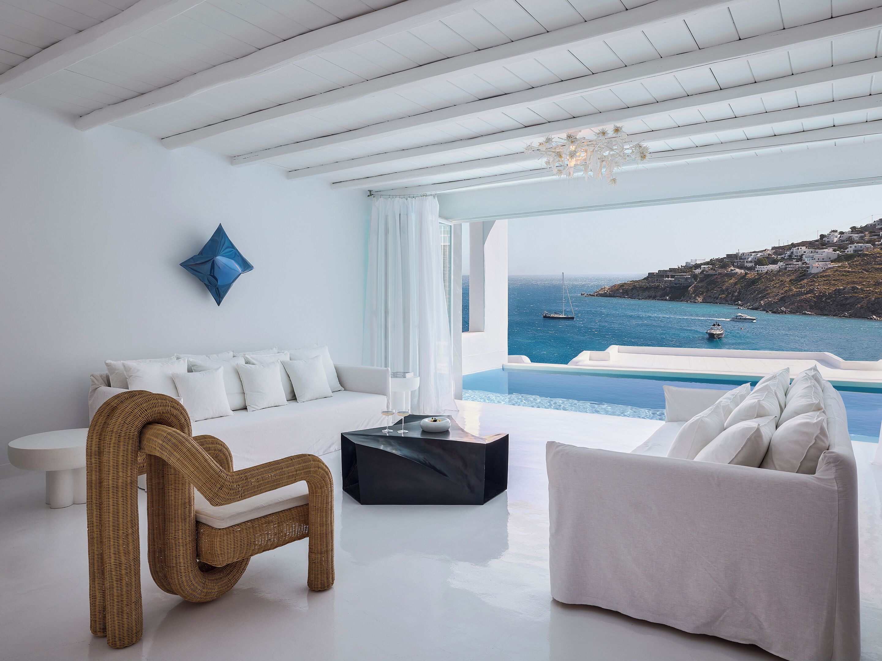 Ein modernes Wohnzimmer mit einem weißen Sofa, einem Korbstuhl und einem schwarzen Couchtisch. Ein großes Fenster bietet Blick auf das Meer und Boote.