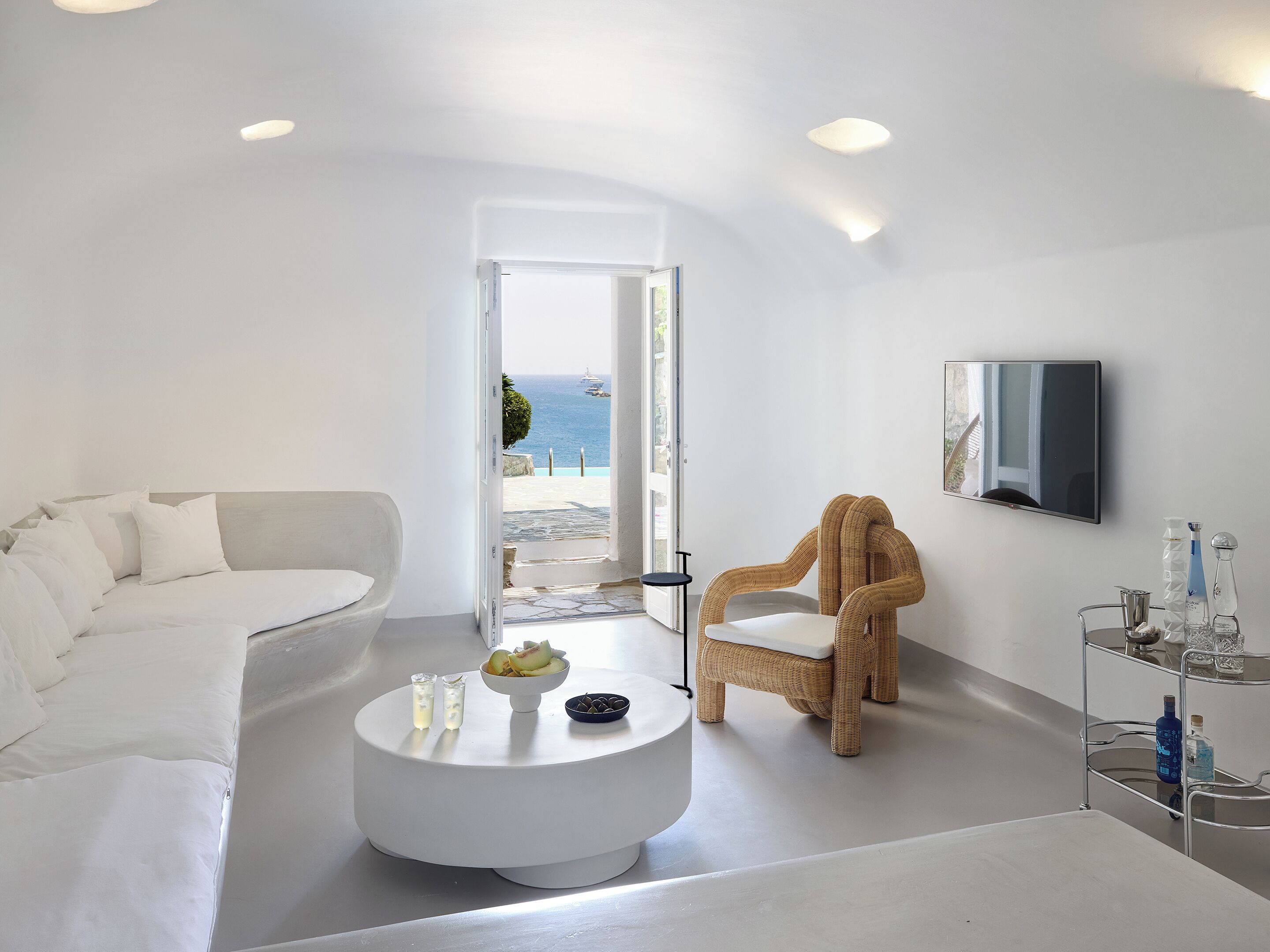 Un salón moderno y minimalista con un sofá blanco, un sillón de mimbre, una mesa redonda de centro con frutas y vasos, y vista al mar a través de una puerta abierta.