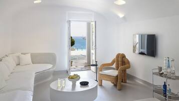 Un salón moderno y minimalista con un sofá blanco, un sillón de mimbre, una mesa redonda de centro con frutas y vasos, y vista al mar a través de una puerta abierta.