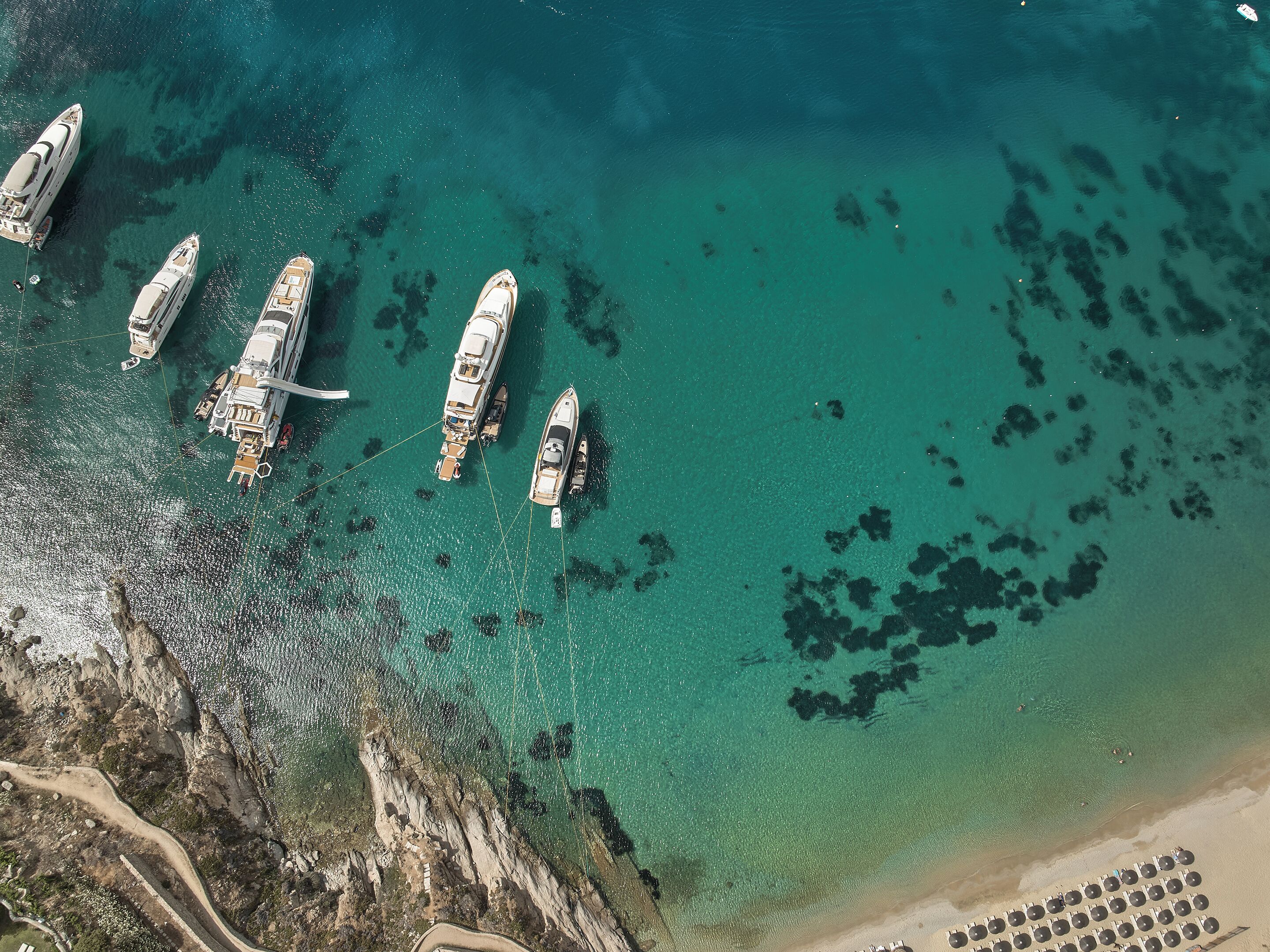 Vista aérea de un puerto deportivo con varios yates amarrados, agua turquesa cristalina y una playa de arena con hileras de sombrillas.