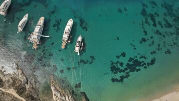 Vista aérea de un puerto deportivo con varios yates amarrados, agua turquesa cristalina y una playa de arena con hileras de sombrillas.