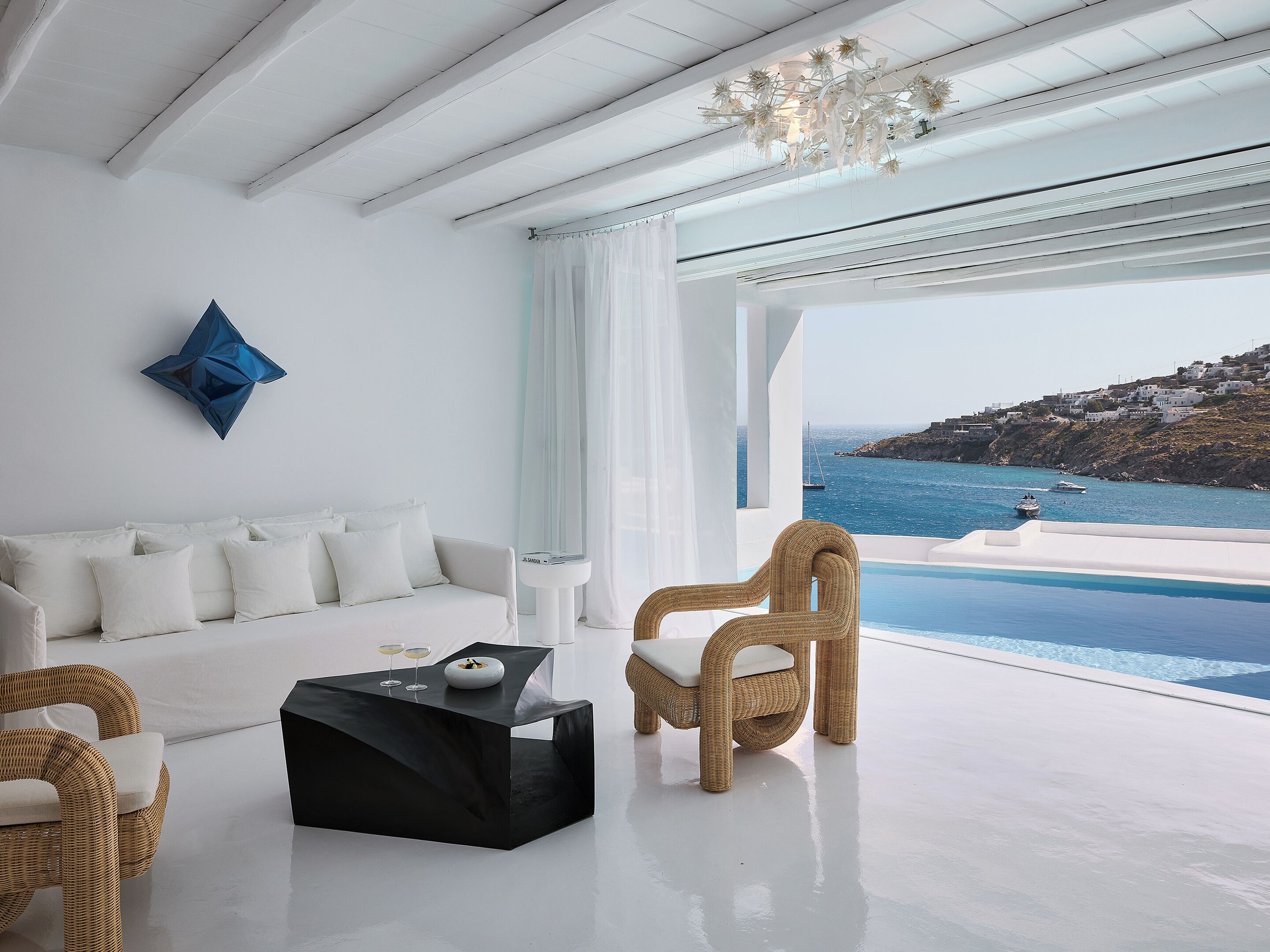 A modern living room with a white sofa, a black coffee table, and a wicker chair. A large window offers a view of the sea and a boat.