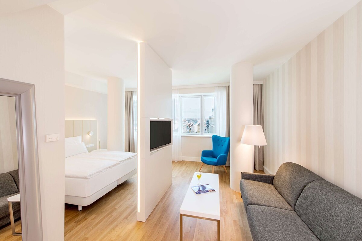 A hotel room with a bed, a TV, a sofa, a chair, a lamp, and a window with a city view.