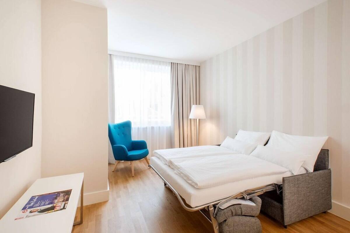 A hotel room with a bed, a TV, a chair, and a window with curtains.