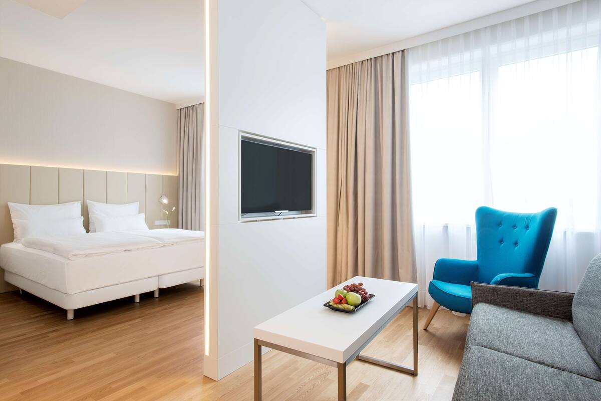 A modern hotel room with a bed, a flat-screen TV, a dining table with fruit, a blue armchair, and a grey sofa.