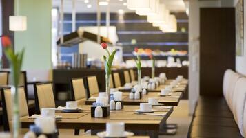 Uma sala de jantar de hotel com mesas compridas dispostas para o serviço, cadeiras e um sofá ao longo da parede.