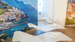 A modern hotel room with a large scenic mural of Amalfi Coast, a white curved bed, a small round table, and a view of the sea. - NH Collection Wien Zentrum (Vienna)