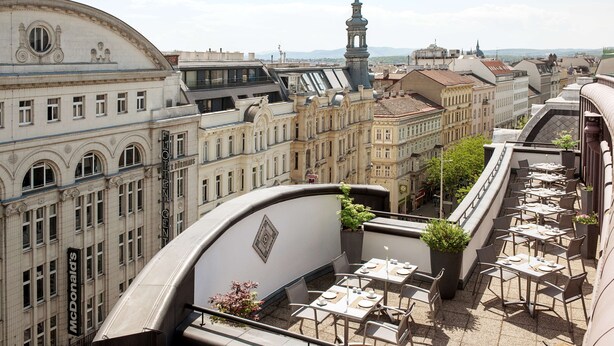 A rooftop terrace with tables and chairs, surrounded by historic buildings with ornate facades and a clock tower in the background. - NH Collection Wien Zentrum (Vienna)