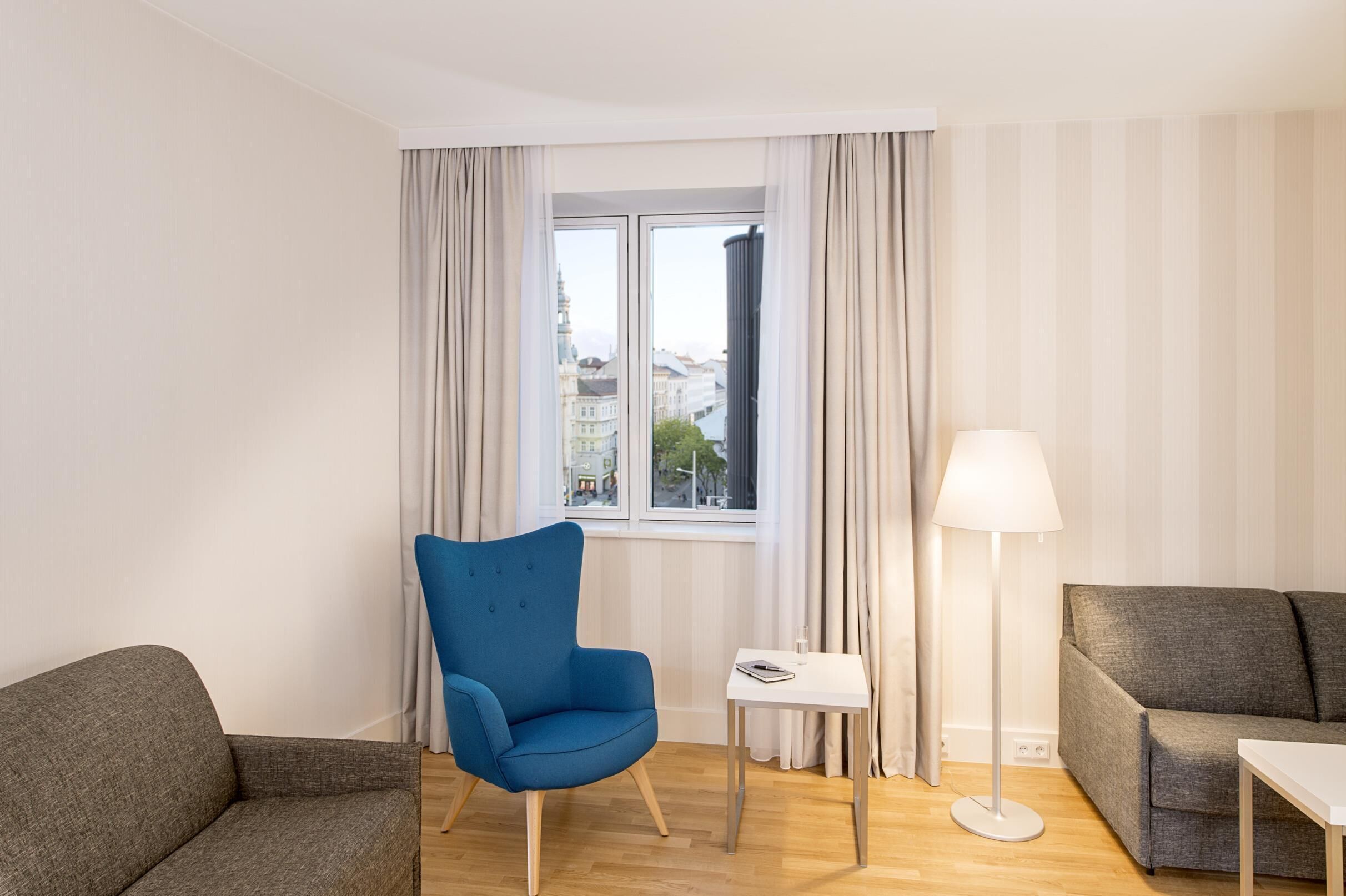 Una sala moderna con un sillón azul, un sofá gris, una mesita blanca y una lámpara de pie. Una ventana con cortinas permite ver edificios al exterior.