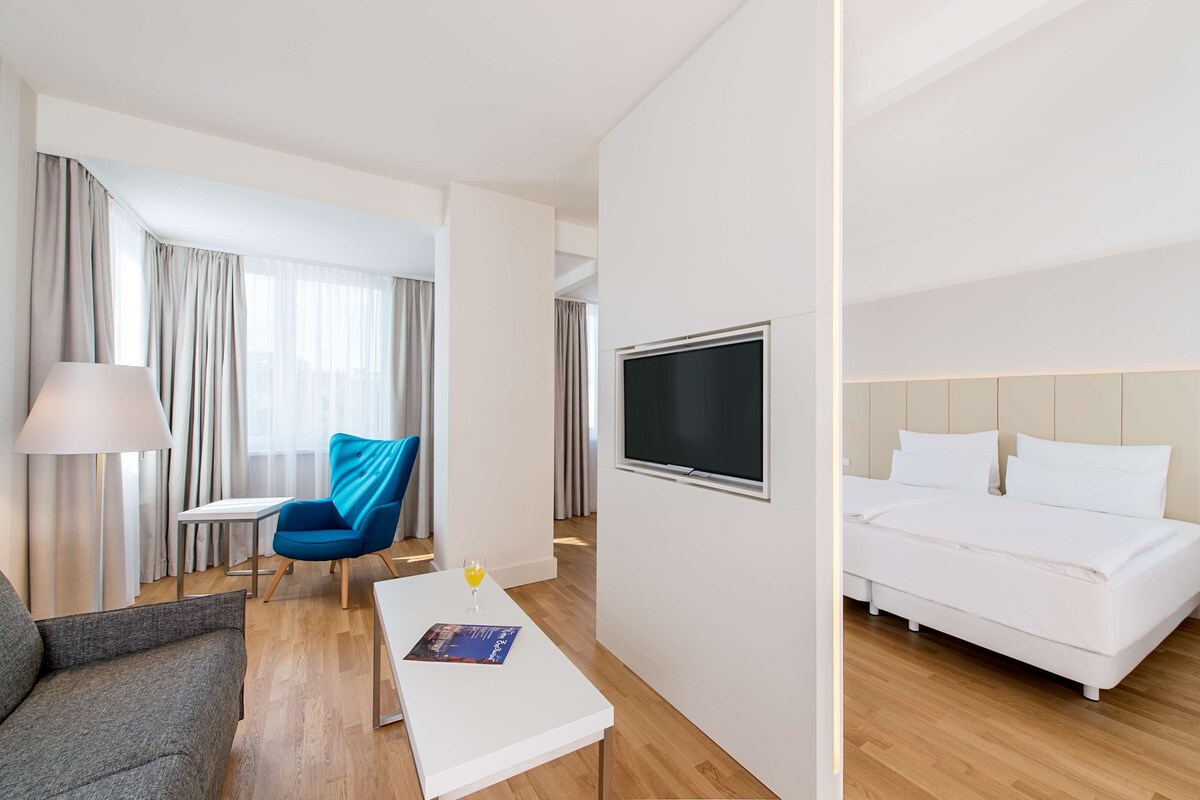 Habitación de hotel con cama, televisión, sofá, sillón, mesa y lámpara.