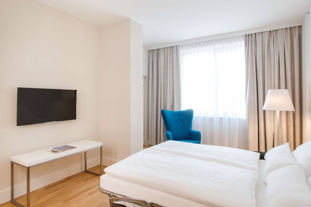 A hotel room with a bed, a TV mounted on the wall, a bedside table, a chair, and a window with curtains.