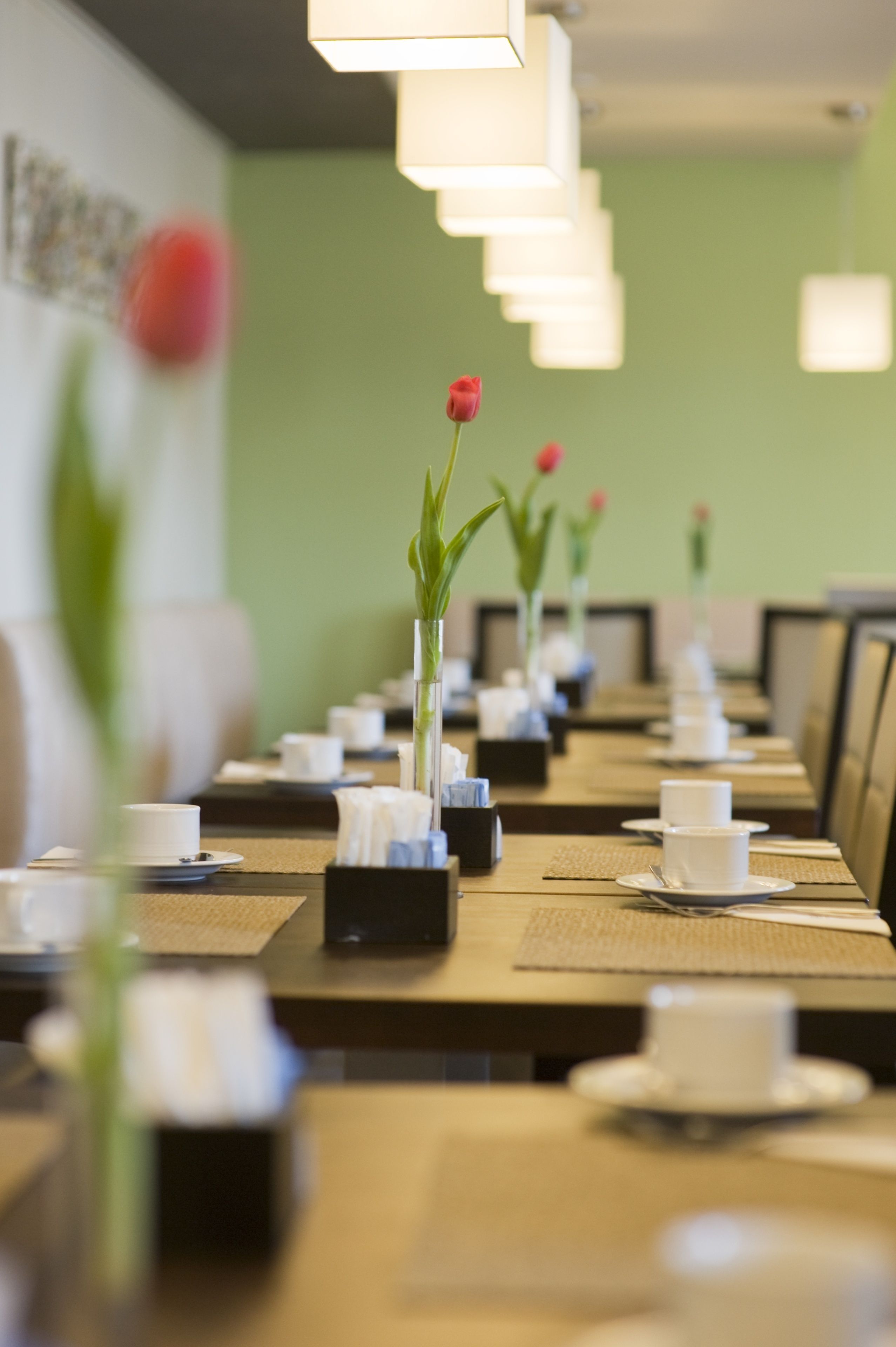 Un restaurante con largas mesas dispuestas para comer, cada una con un pequeño jarrón de flores y vajilla ordenada.