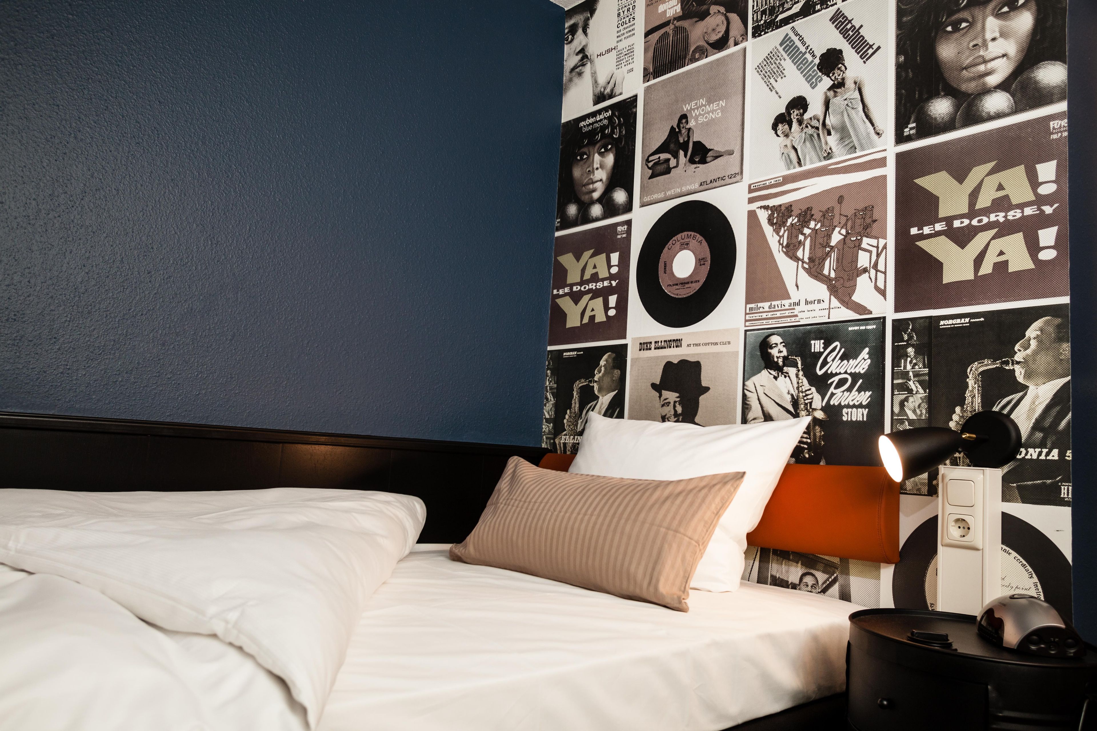 A bedroom with a bed, pillows, a nightstand, and a wall covered with black and white photographs and text.