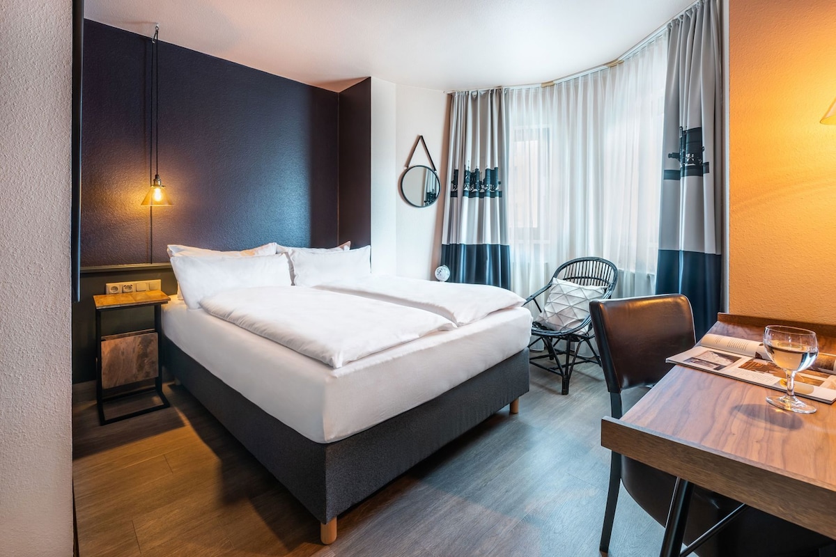 Habitación de hotel con cama, escritorio con silla, mesa con vaso de vino y ventana con cortinas.