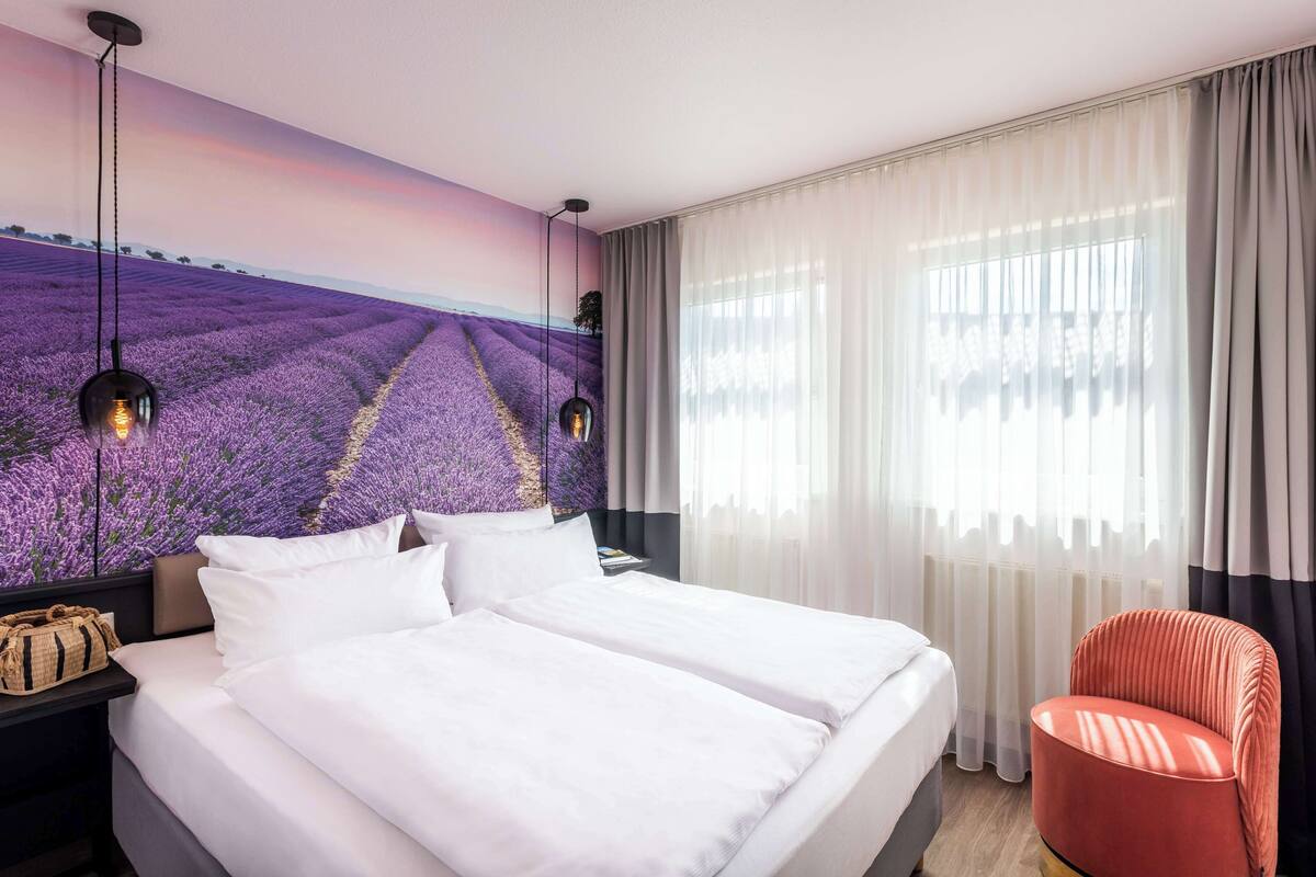 A hotel room with a large bed, a red chair, a nightstand with a lamp, and a wall mural of lavender fields.
